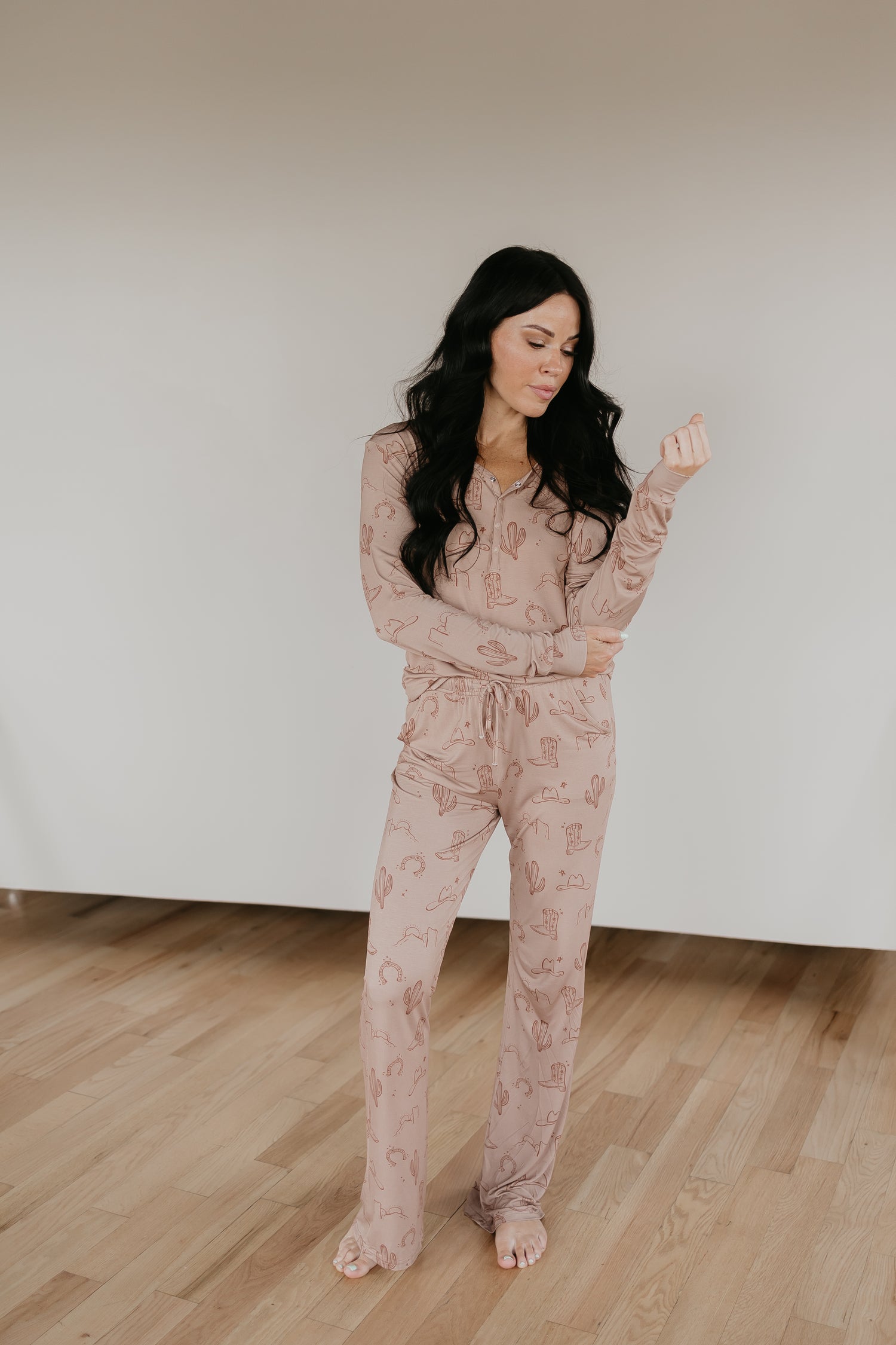 A woman with long, dark hair is standing barefoot on a wooden floor. She is wearing Women's Bamboo Pajamas from the Minty x FF Wild West Desert Dreams collection, made from breathable, hypo-allergenic fabric featuring a cactus and geometric pattern. With a playful expression, she holds her left sleeve with her right hand against the plain, light-colored background.