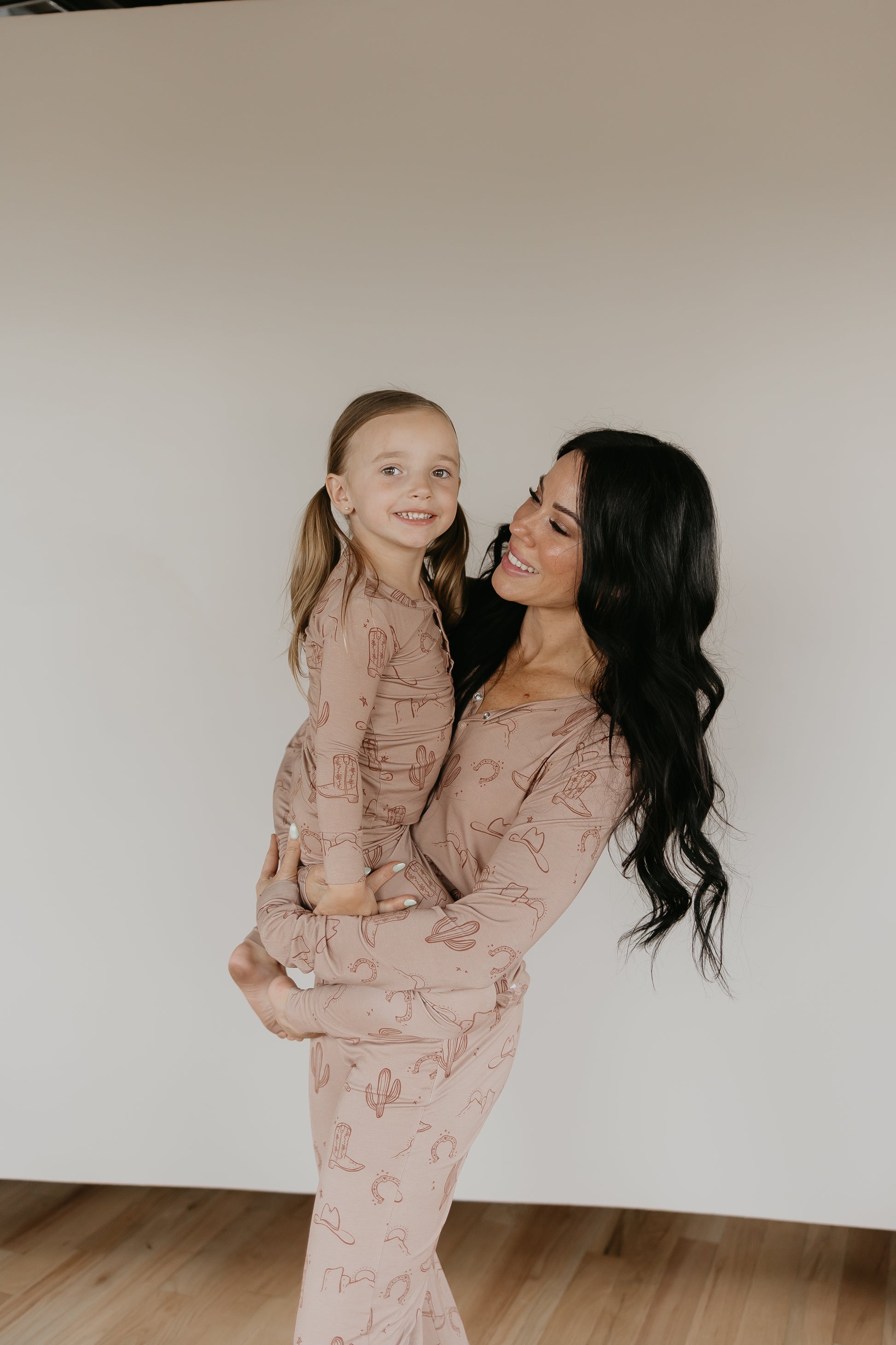 A woman with long, dark hair holds a young child. Both are smiling and wearing matching Women's Bamboo Pajamas from the Minty x FF Wild West Desert Dreams collection in a playful cactus and desert theme made of breathable fabric. They stand on a light wooden floor against a plain backdrop.