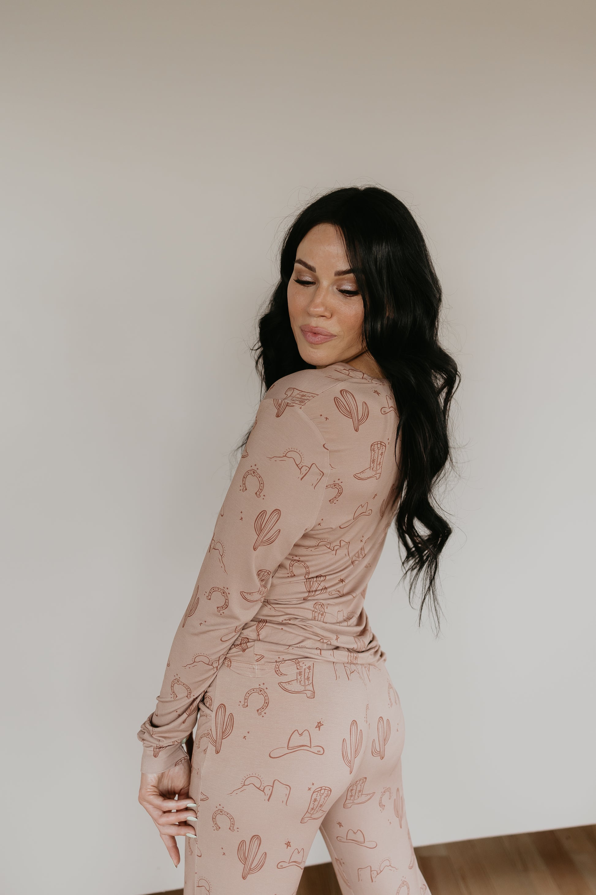 A woman with long dark hair stands against a light background, wearing the Women's Bamboo Pajamas from the Minty x FF Wild West Desert Dreams collection. The matching beige loungewear features a cactus print and is made from breathable fabric. She looks down over her shoulder with a serene expression.