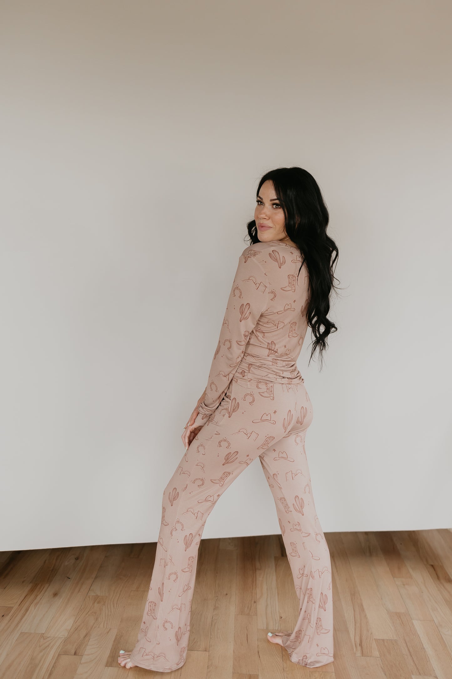 A woman with long dark hair stands barefoot indoors on a wooden floor, wearing a beige Women's Bamboo Pajama set with pink patterns from Minty x FF Wild West Desert Dreams. She is turned slightly to the side and looking over her shoulder toward the camera.