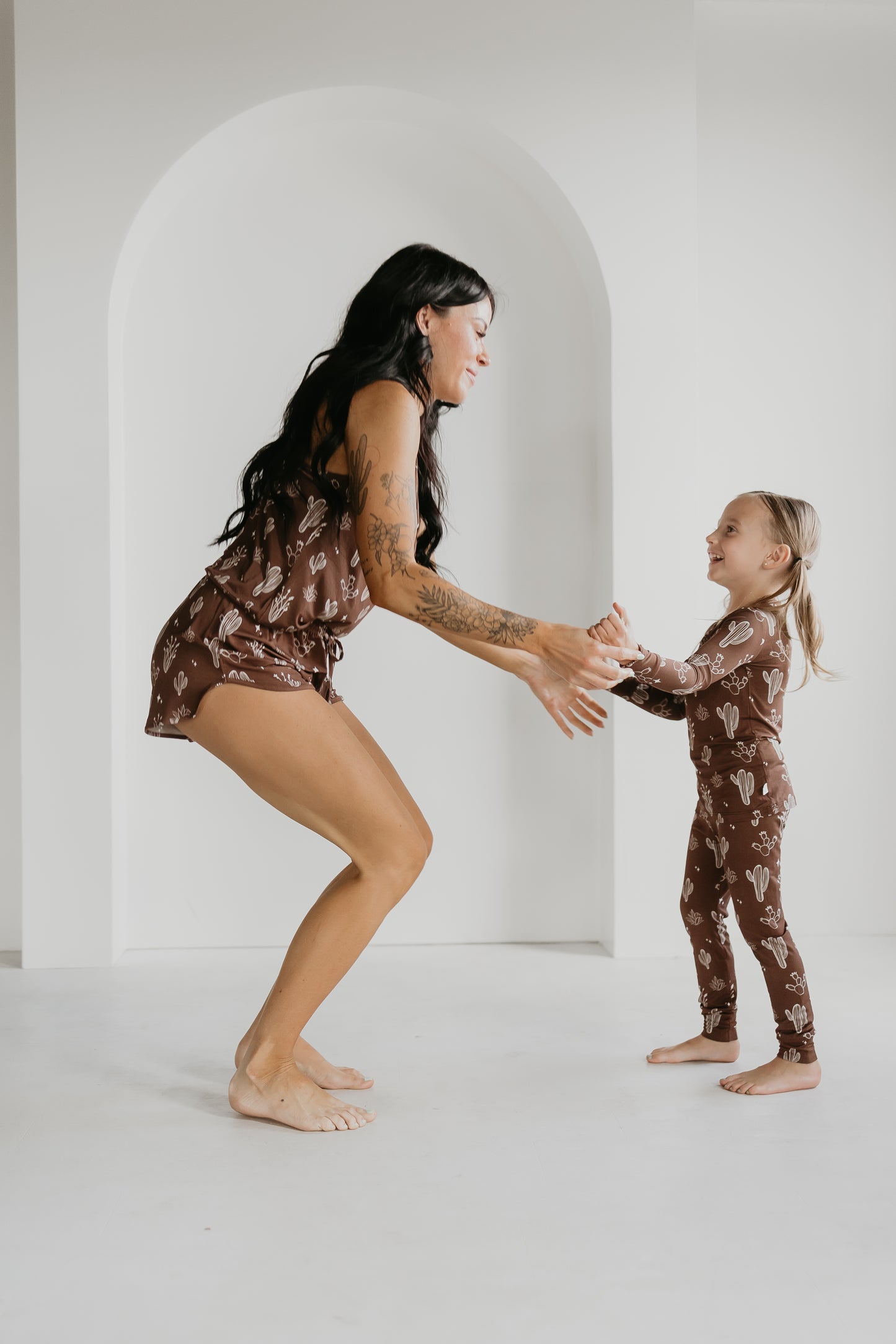 A woman and a young girl, both wearing matching brown outfits with cactus patterns from the Minty x FF Wild West Desert Dreams collection, smile at each other in a brightly lit room. The woman is bending slightly with her arms extended toward the girl, who reaches out with both hands.