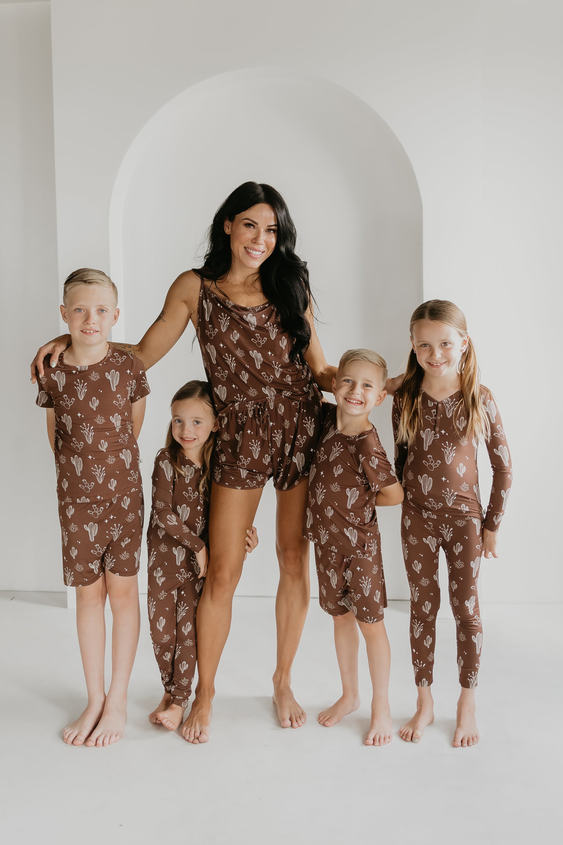 A woman and four children are gathered together, smiling in their coordinated brown Bamboo Two Piece Pajamas with white patterns from the Minty x FF Wild West Desert Dreams collection. They stand barefoot against a well-lit room with a white arched backdrop.