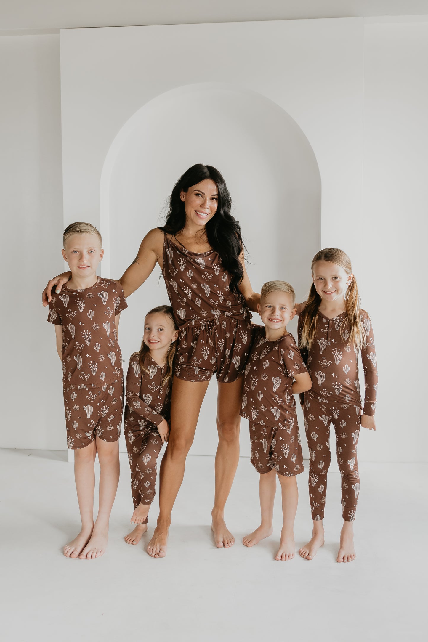 A woman and four children stand barefoot in matching brown floral outfits from the Minty x FF Wild West Desert Dreams collection against a minimalist white background. They are smiling, with the woman in the center and two children on each side.