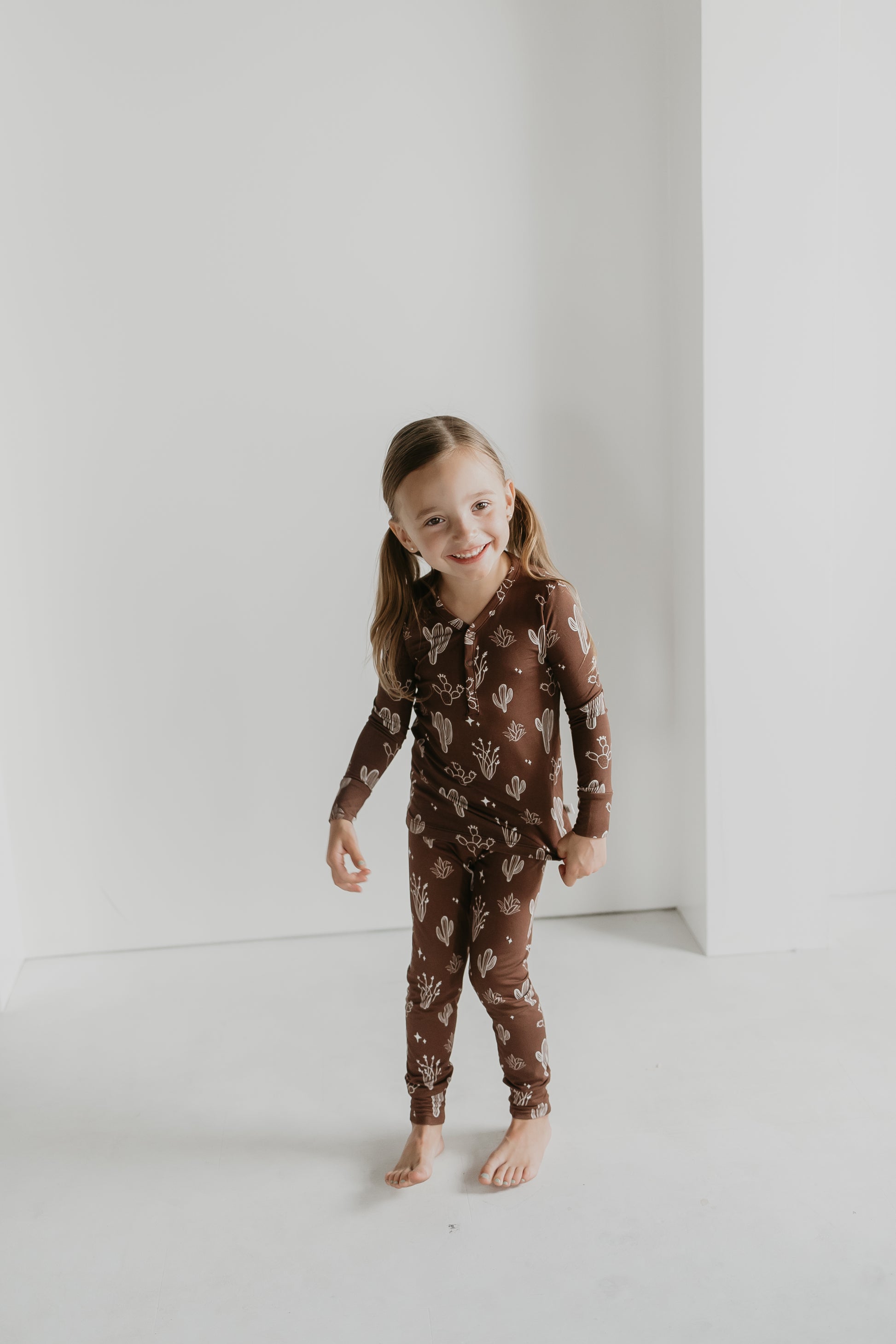 A young girl stands barefoot on a light-colored floor against a plain white wall, showcasing the Minty x FF Wild West Desert Dreams collection. She smiles in her brown, floral pattern long-sleeved Bamboo Two Piece Pajamas from the Minty x ff Desert Dreams line, with her hair styled in two ponytails.