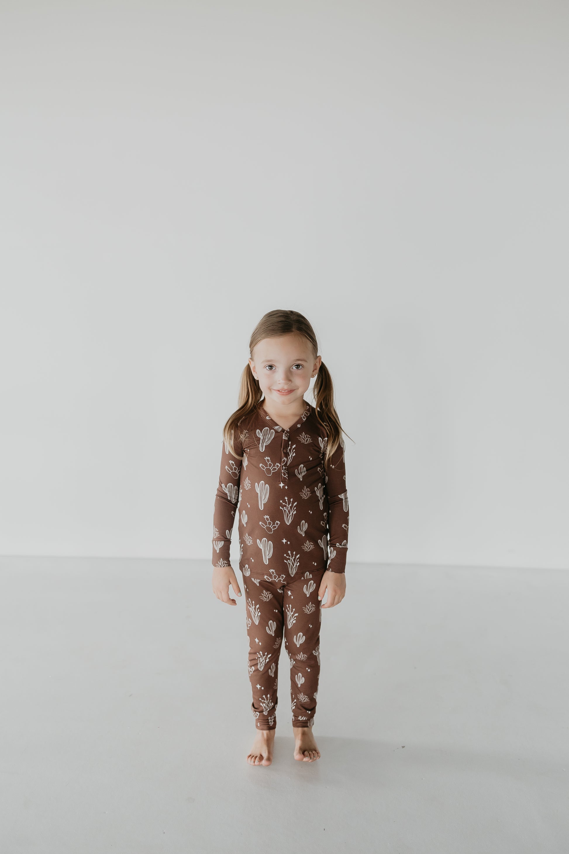 A young child with long hair stands barefoot in Bamboo Two Piece Pajamas from the Minty x FF Wild West Desert Dreams collection, showcasing a brown set adorned with white cactus patterns. She smiles gently against a plain, light-colored background.