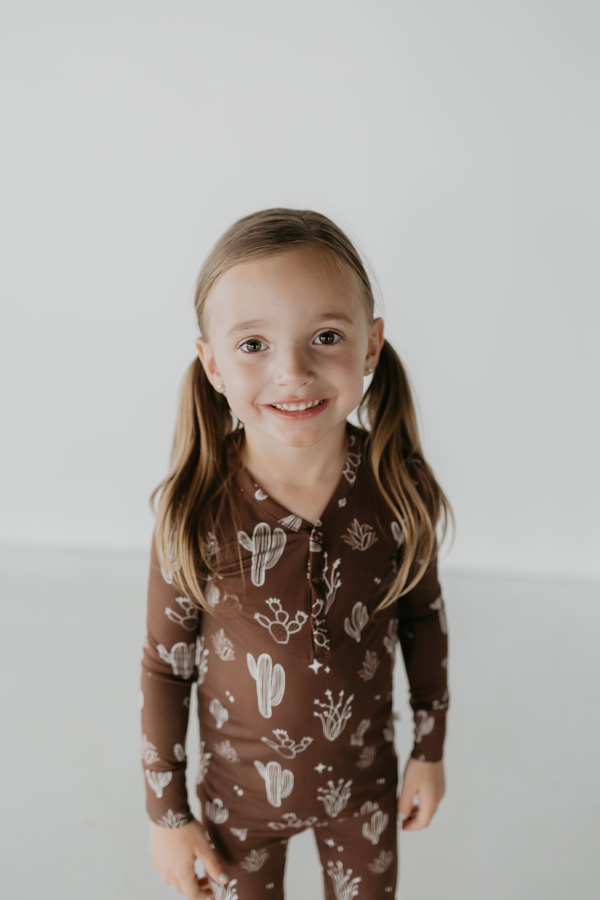 A young girl with long hair in pigtails smiles at the camera, showcasing her brown outfit adorned with white cactus and plant illustrations from the Minty x FF Wild West Desert Dreams collection. The hypo-allergenic Bamboo Two Piece Pajamas complement the simple light gray background perfectly.