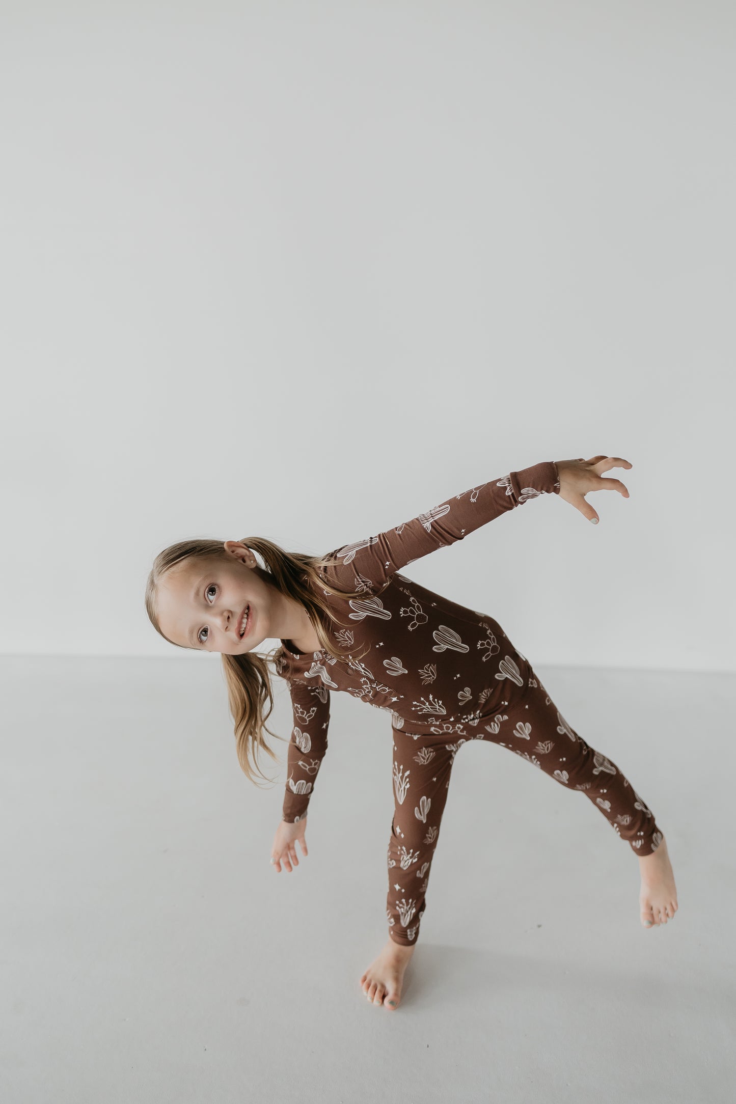 A young girl in the Minty x ff Desert Dreams Bamboo Two Piece Pajamas cheerfully balances on one foot against a plain white background. Her light brown hair cascades as she smiles, enjoying the comfort and style of her hypoallergenic clothing from the Minty x FF Wild West Desert Dreams collection.