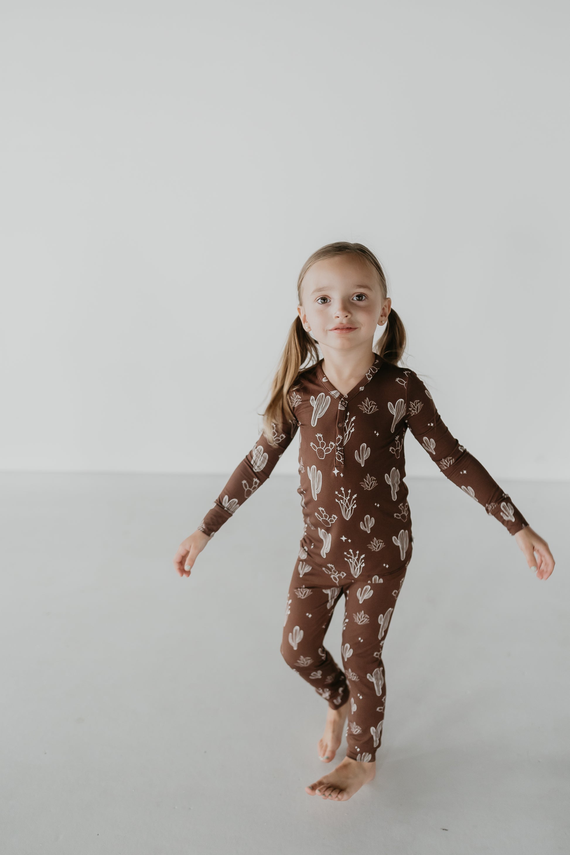 A young child with pigtails walks barefoot on a light-colored floor, dressed in charming bamboo two piece pajamas featuring a cactus pattern from the Minty x FF Wild West Desert Dreams collection. The hypo-allergenic pajamas ensure comfort against the backdrop of a plain white wall.