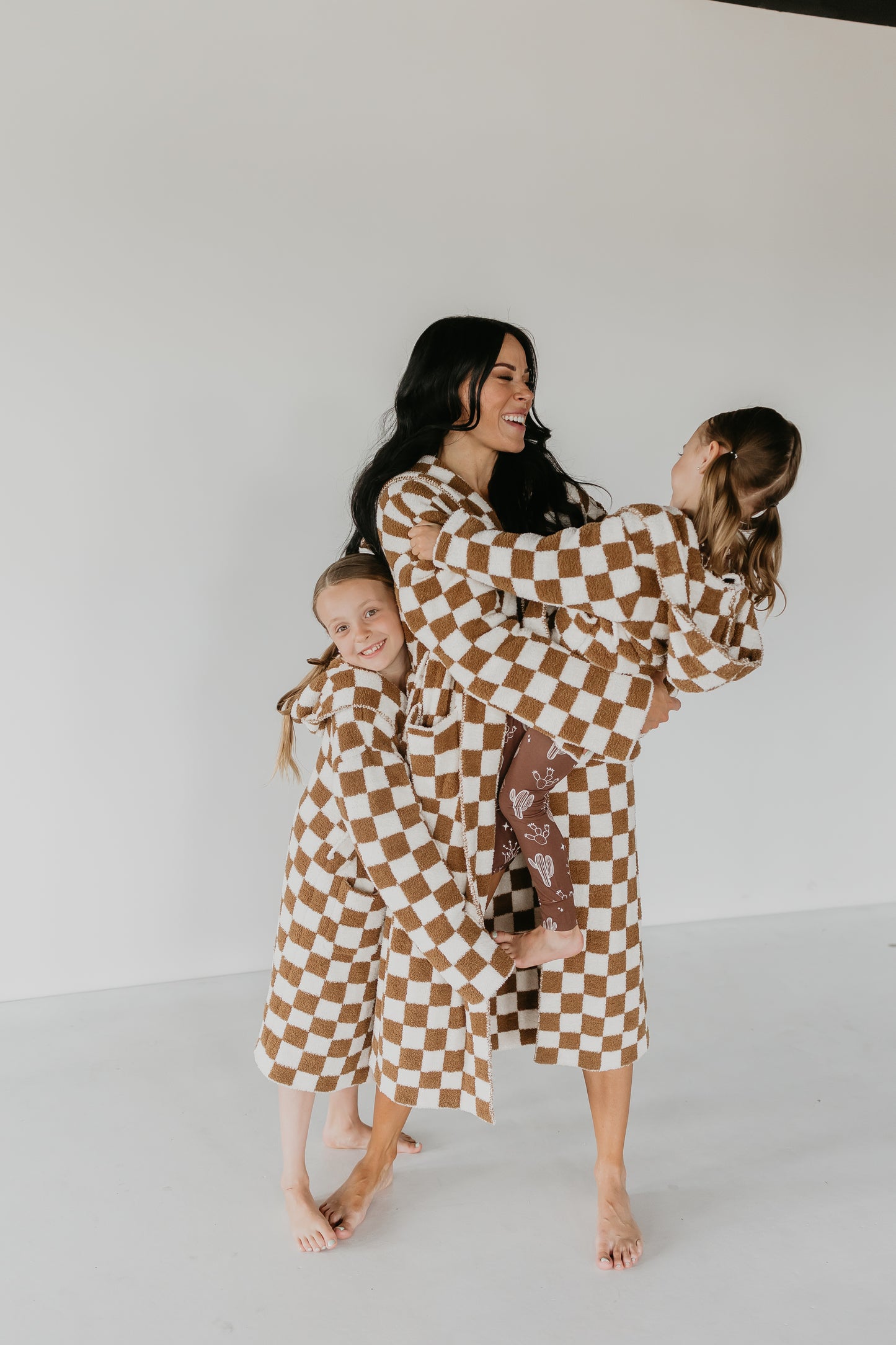 A woman and two girls, all dressed in matching Adult Hooded Robes from the Minty x FF Wild West Desert Dreams collection, share a joyful moment. The girls stand on either side of the woman, holding onto her affectionately. They are barefoot against a plain white background.