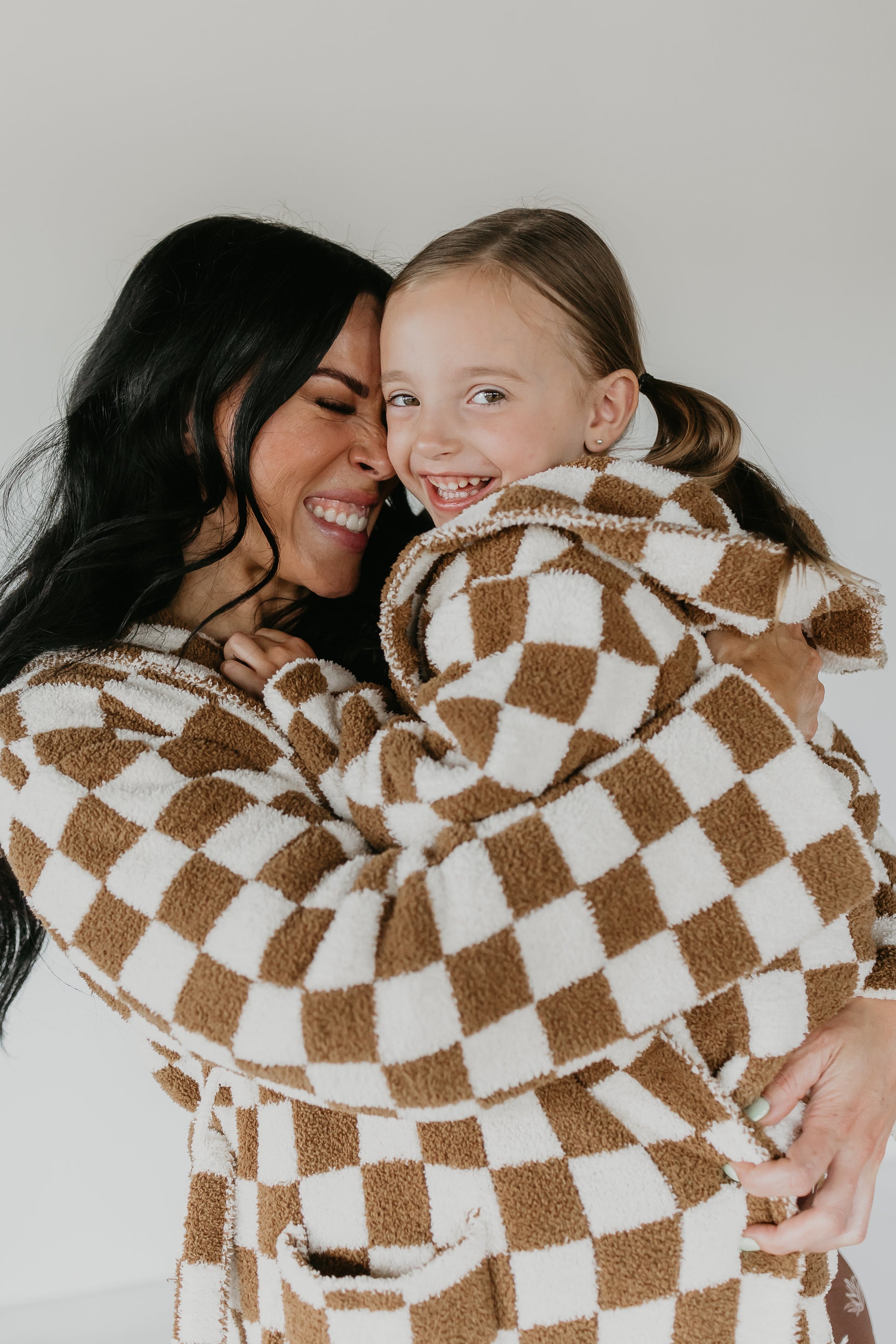 A woman and a young girl, both wrapped in the luxurious Minty x FF Wild West Desert Dreams Adult Hooded Robe in a brown and white checkered design, are embracing and smiling. The woman, with long dark hair, shares this joyful moment with the girl who has light brown hair tied back. Both exude happiness and contentment.