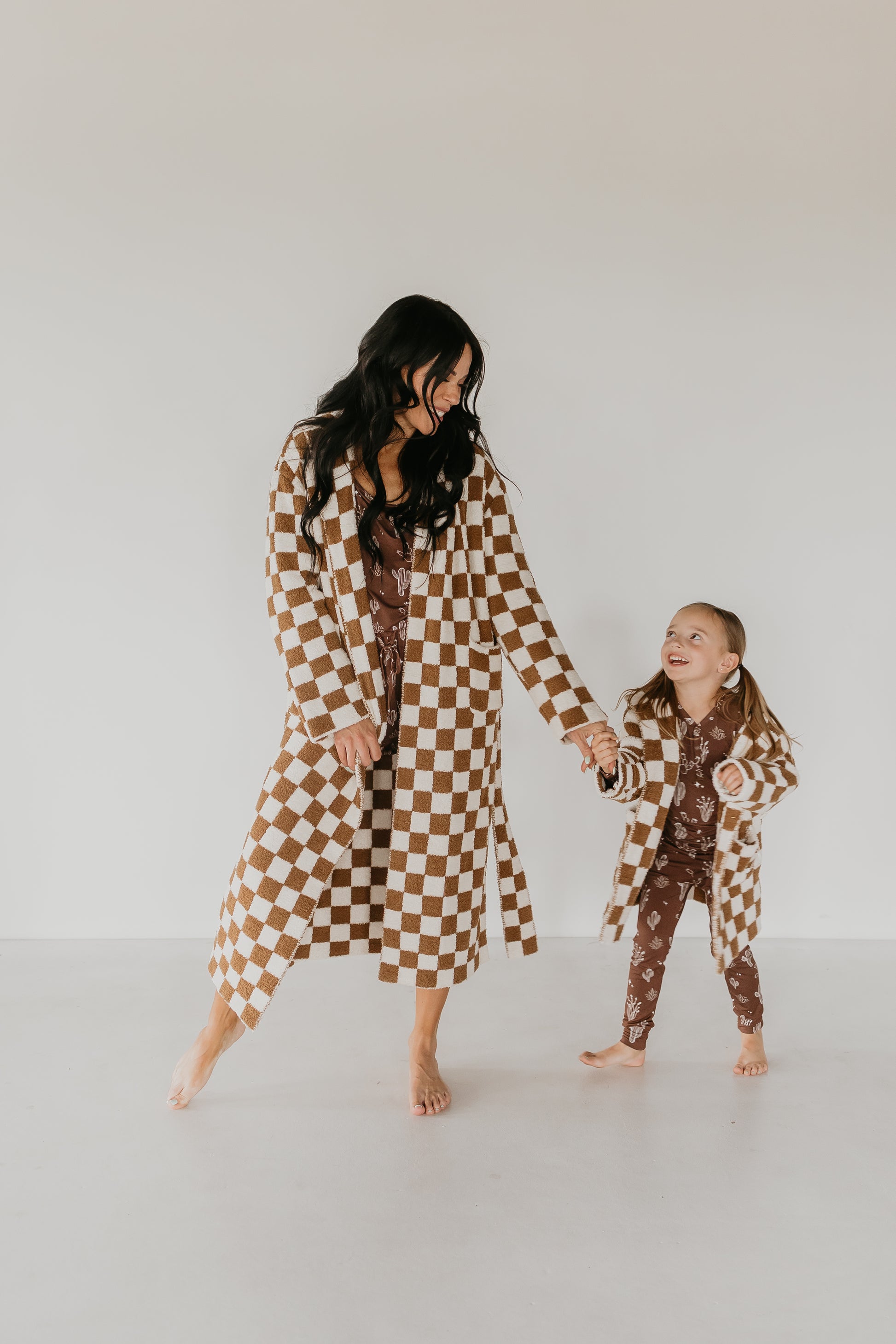 A woman and a young girl sport matching robes from Minty x FF Wild West Desert Dreams, showcasing their stylish Adult Hooded Robes in brown and white checks complemented by patterned outfits. Smiling as they hold hands in a minimalist, neutral-colored space, they enjoy the comfort and safety of Oeko-tex certified materials.