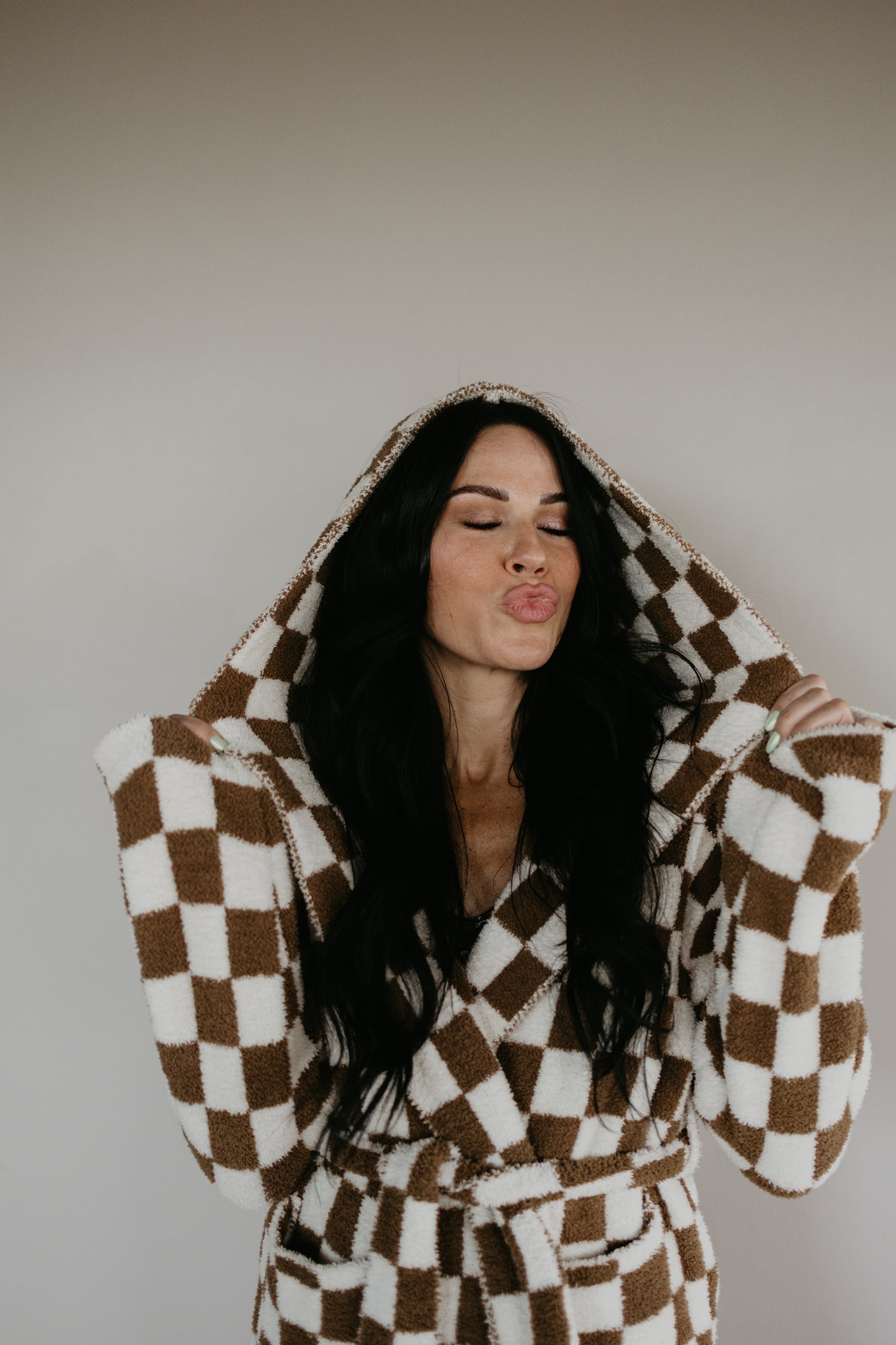 A person with long dark hair is wearing an Adult Hooded Robe from Minty x FF Wild West Desert Dreams, crafted from microfiber feather yarn. They are making a playful expression with closed eyes and puckered lips. The robe, Oeko-tex certified for comfort, stands out against the plain background.