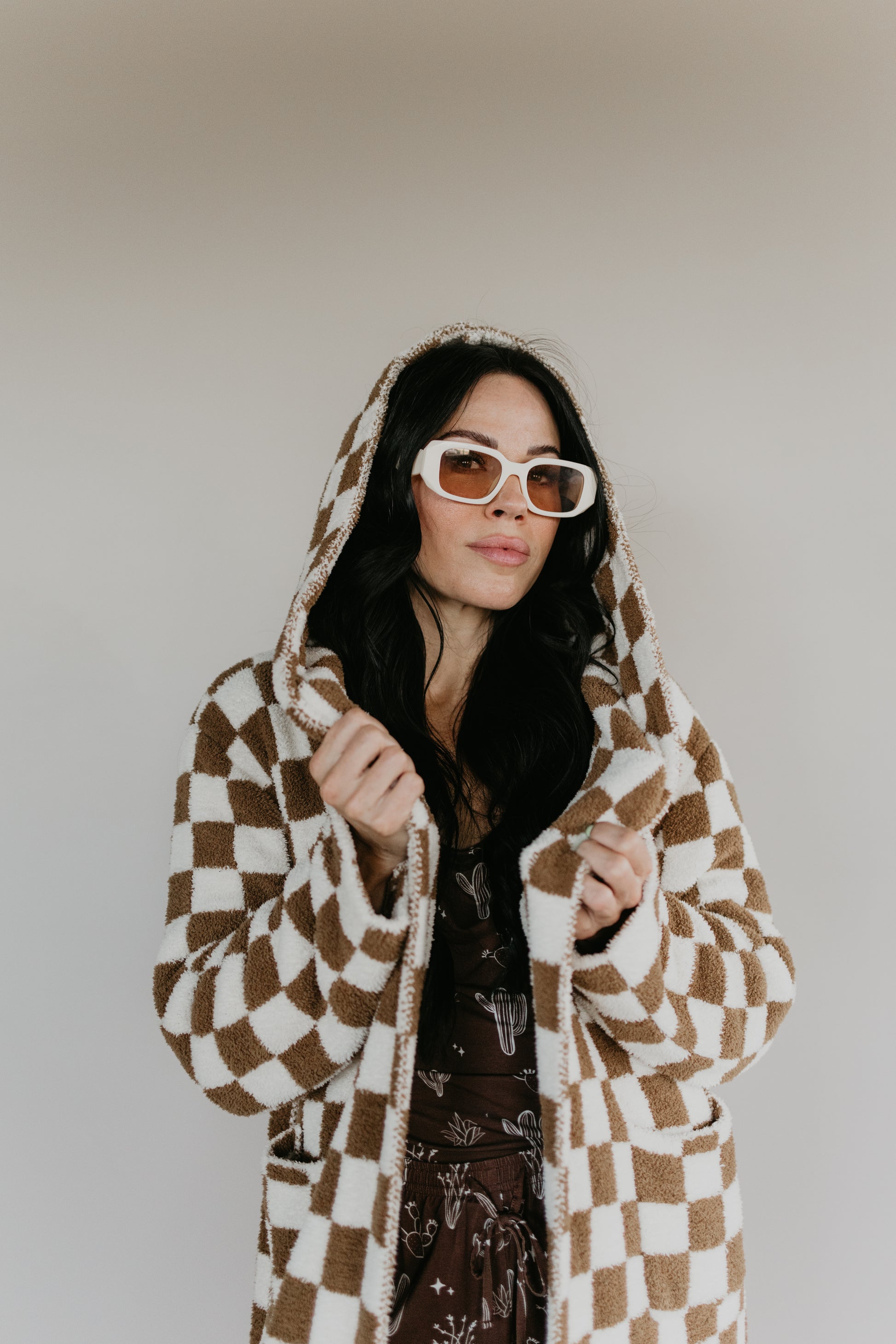 A person with long dark hair, sporting oversized white sunglasses and an Adult Hooded Robe from the Minty x FF Wild West Desert Dreams collection, featuring a brown and white checkered pattern crafted from microfiber feather yarn, stands against a neutral background.