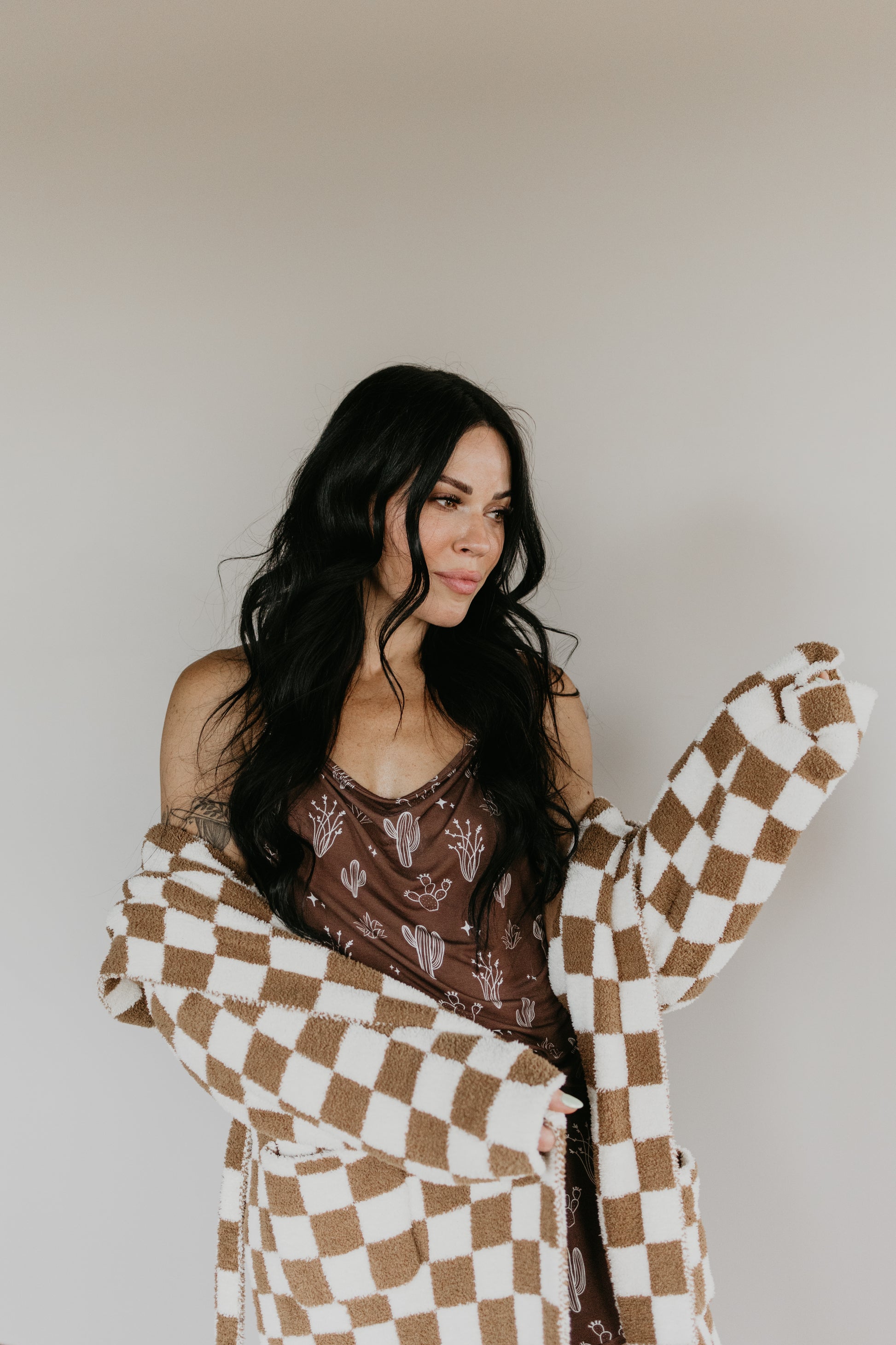 A woman with long black hair smiles gently as she stands against a plain background, wearing a sleeveless brown top with a cactus design. She drapes an Oeko-tex certificated cozy brown and white checkered Adult Hooded Robe from the Minty x FF Wild West Desert Dreams collection over her shoulders.