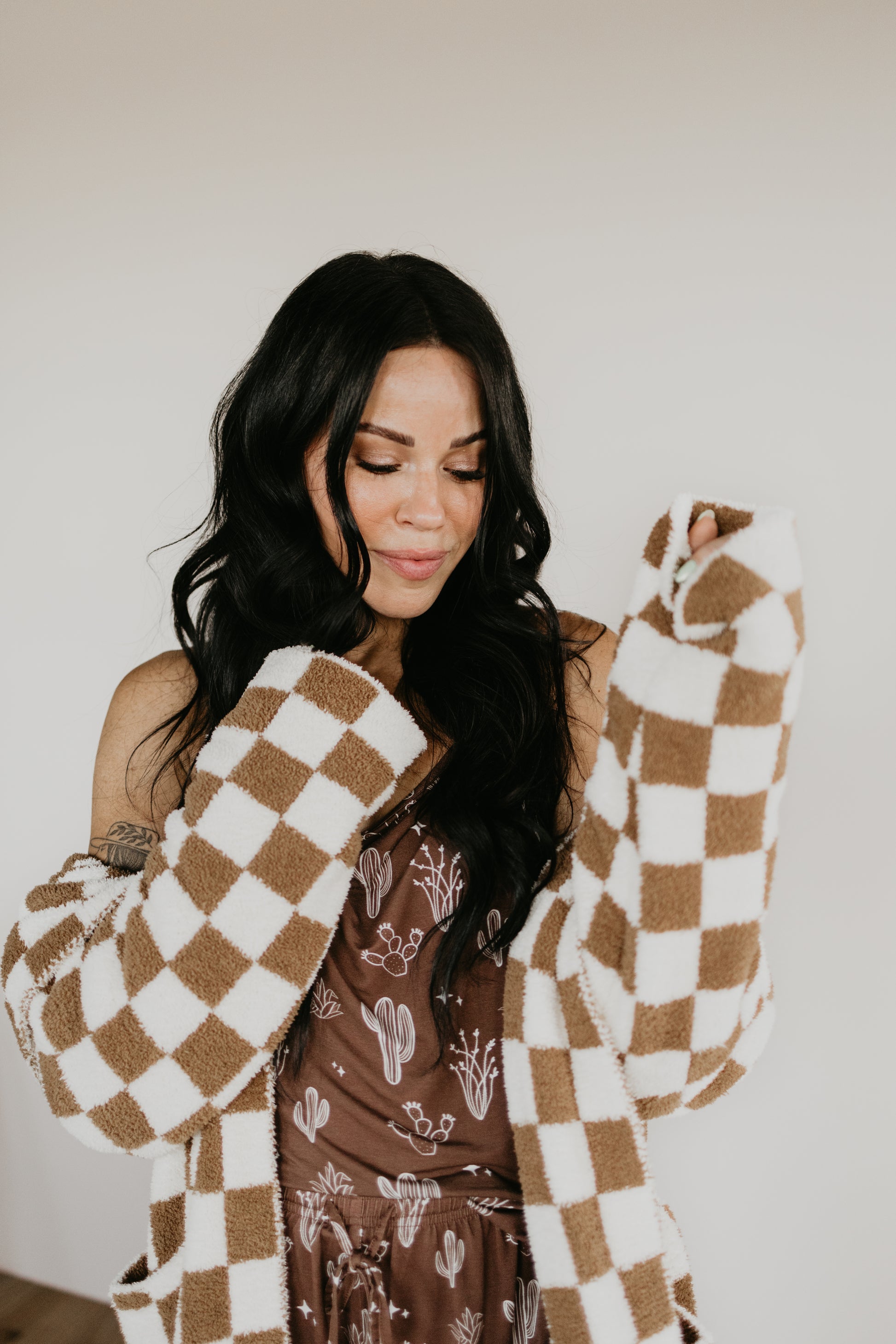 A woman with long dark hair wearing an Adult Hooded Robe by Minty x FF Wild West Desert Dreams, featuring a brown pattern and a cozy checkered jacket made of microfiber feather yarn. Her eyes are closed as she gently holds the velvety-rich texture. The simple, neutral background enhances her relaxed pose.