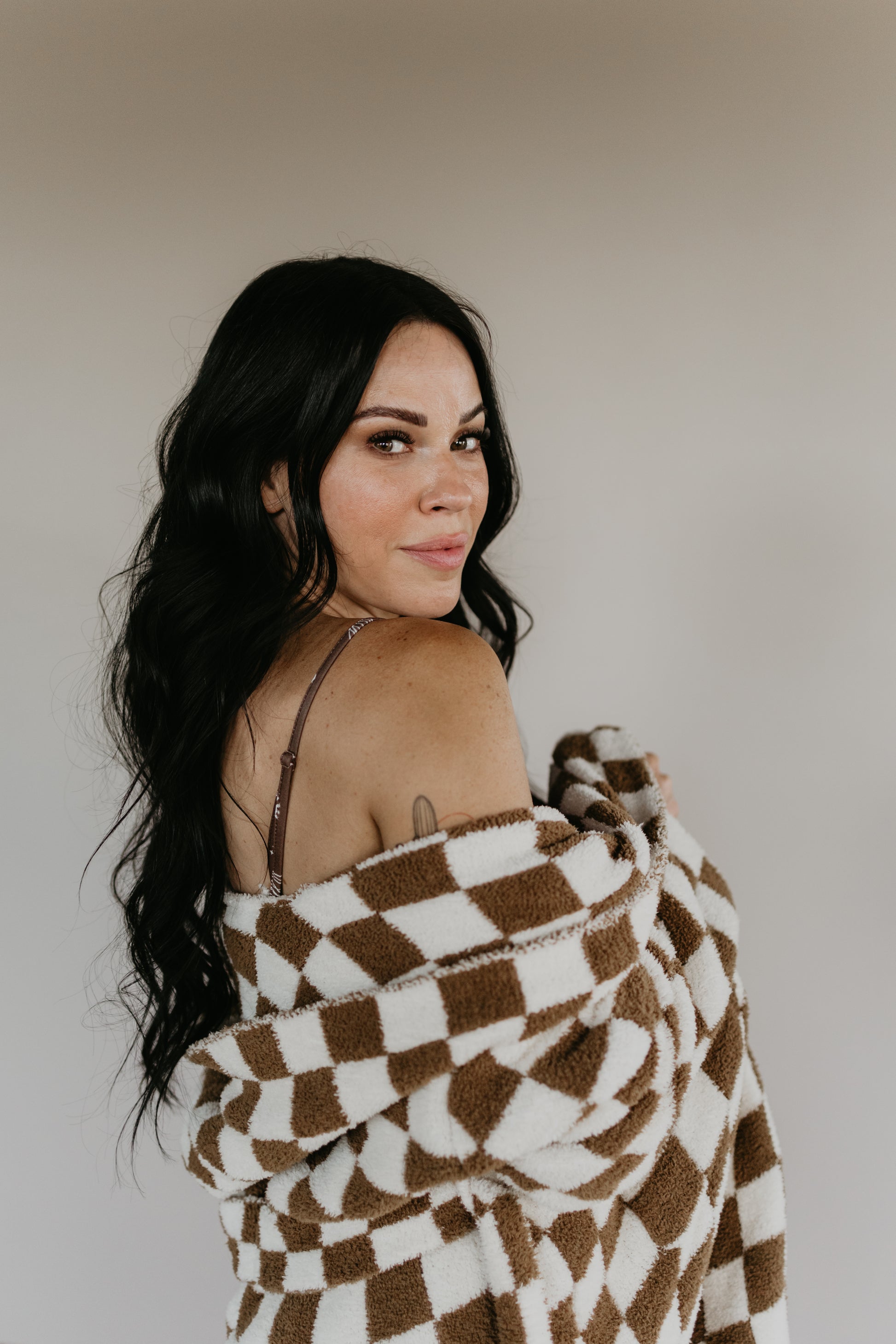 A woman with long dark hair glances over her shoulder, draped in the luxurious texture of a brown and white checkered Adult Hooded Robe by Minty x FF Wild West Desert Dreams. She stands against a neutral background, cocooned in the velvety embrace of the robe.