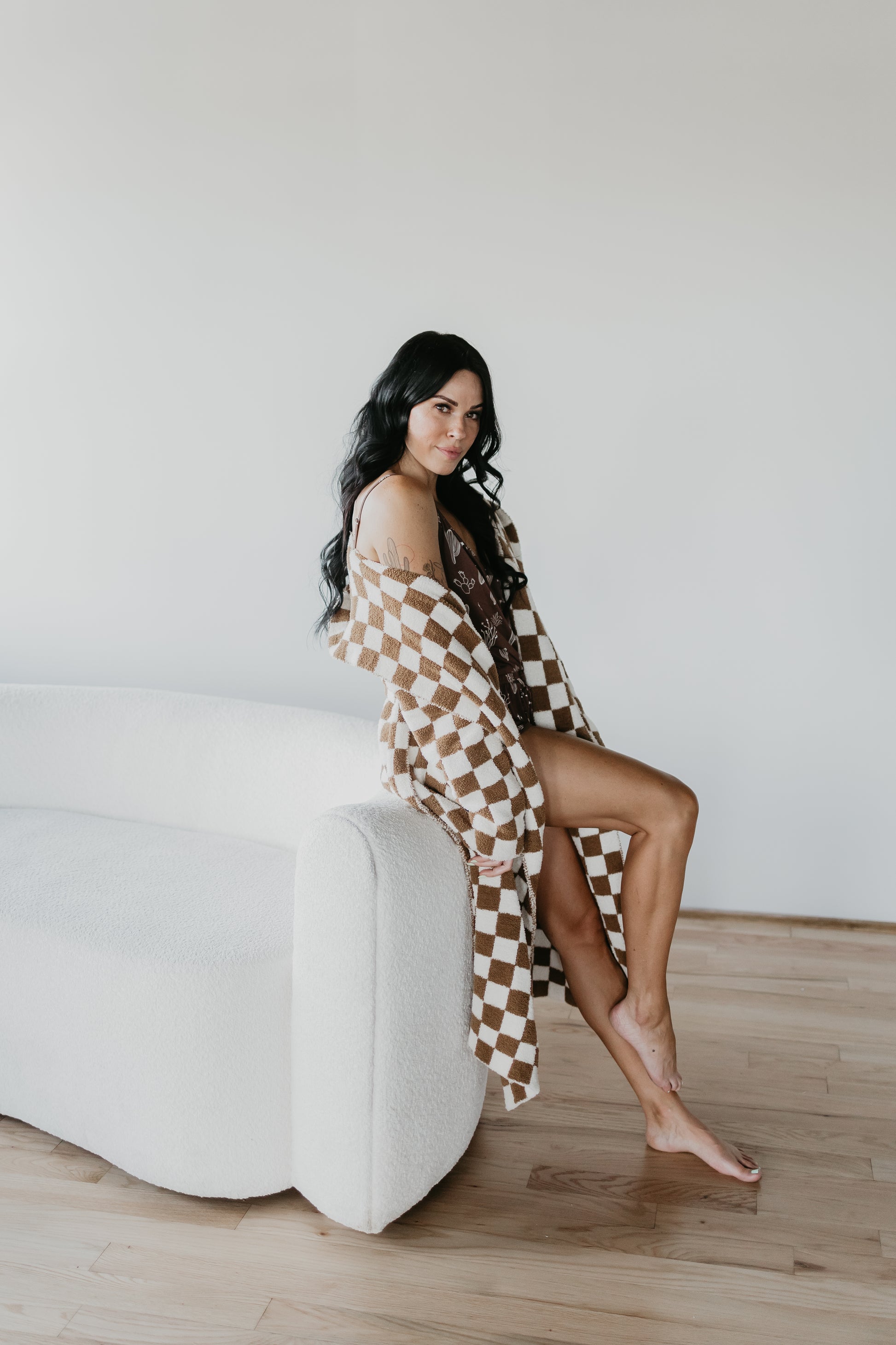 A woman with long, dark hair sits on the armrest of a white couch, draped in an Oeko-Tex certified Adult Hooded Robe from the Minty x FF Wild West Desert Dreams collection. She displays her bare legs against a backdrop of light wood flooring and a plain white wall, embodying the timeless ambiance of Forever French Baby.