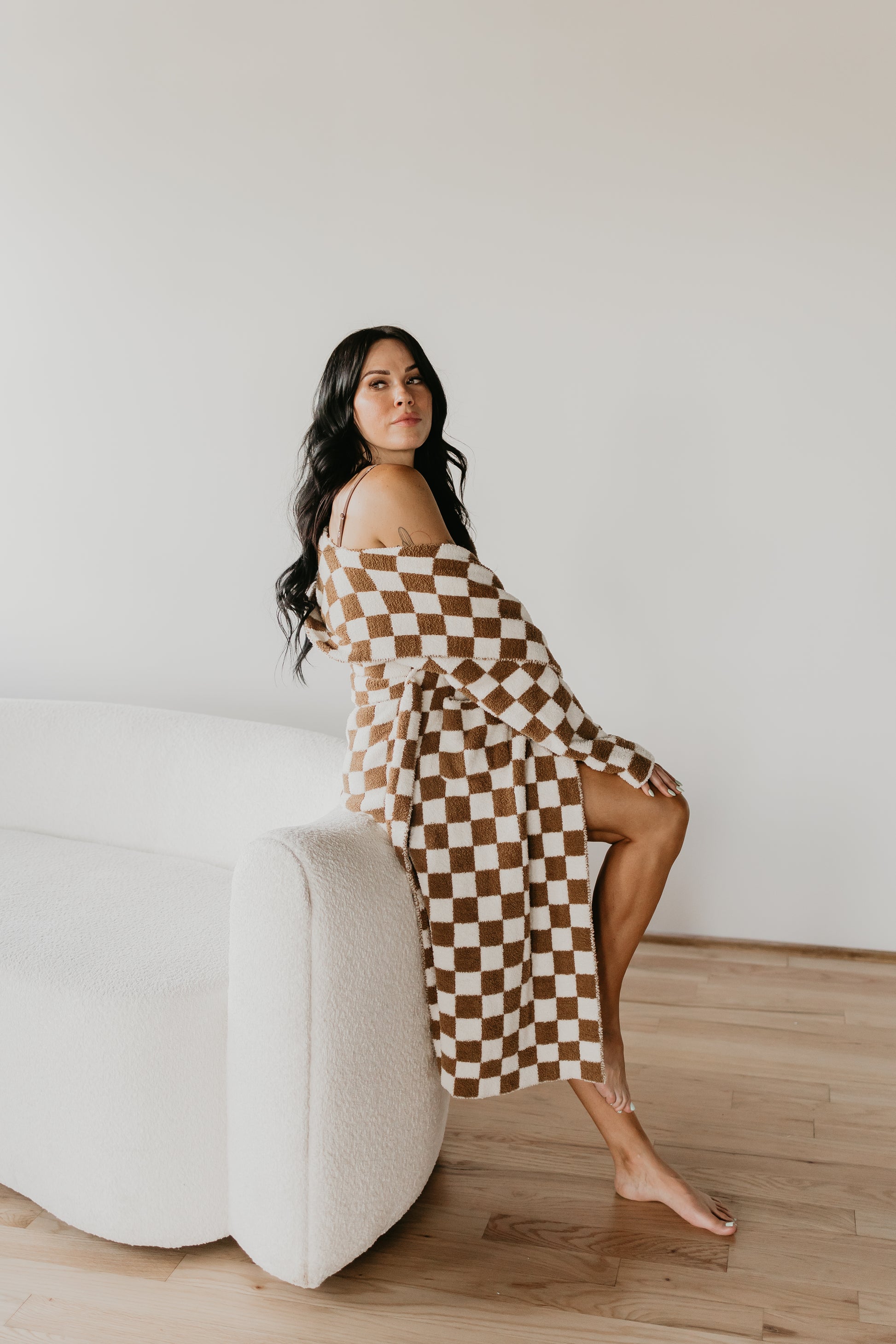 A woman with long dark hair sits on the arm of a white sofa, wearing an off-the-shoulder Adult Hooded Robe by Minty x FF Wild West Desert Dreams. The robe features a brown and white checkered pattern and is crafted from luxe microfiber feather yarn. She is barefoot on a wooden floor against a plain white wall backdrop.