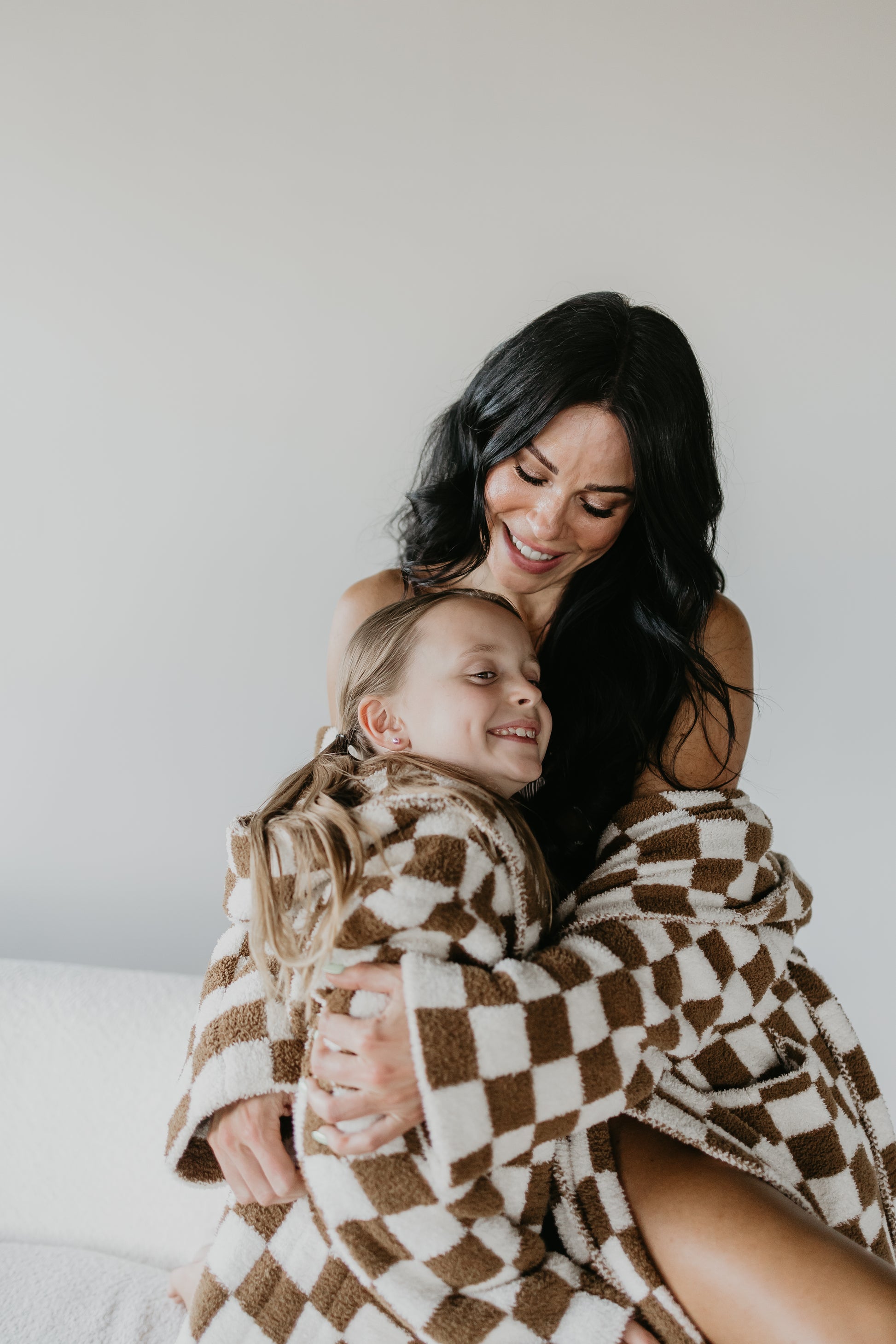 A woman and a young girl are sitting together, both smiling warmly as they enjoy their Minty x FF Wild West Desert Dreams Adult Hooded Robe in the checkered pattern. The Oeko-Tex certified materials wrap them in cozy comfort, while the softly lit background adds to the calm and joyful atmosphere.