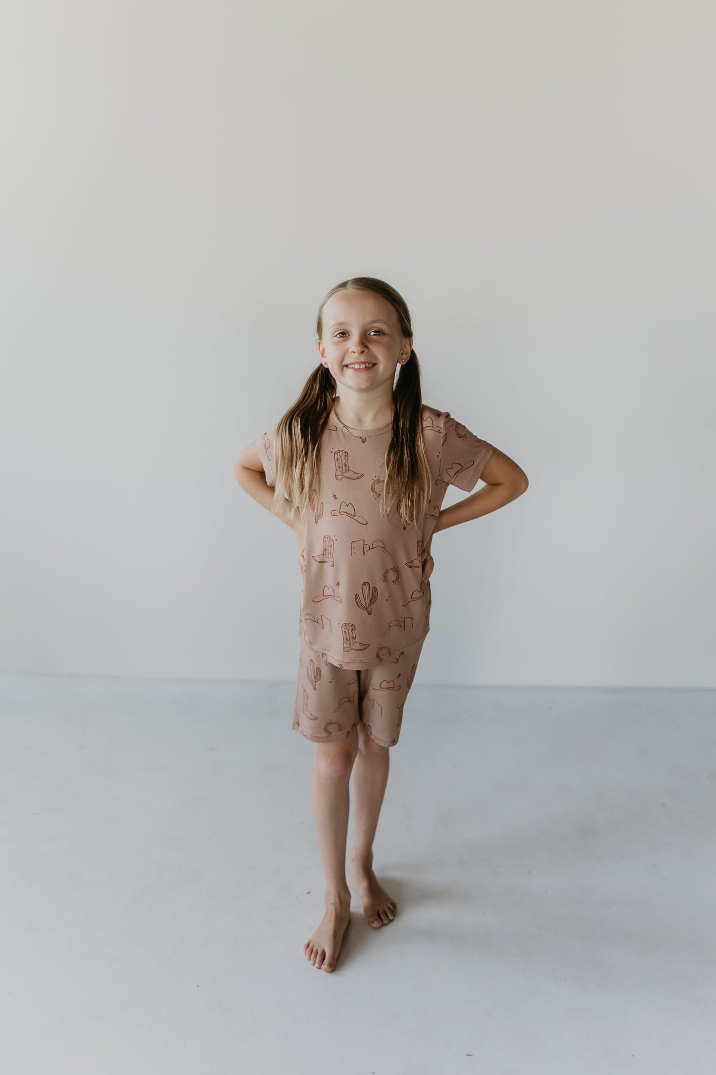 A young girl stands barefoot against a plain white background. She is wearing the Bamboo Short Set from Minty x FF Wild West Desert Dreams, featuring a brown shirt and matching shorts made of eco-friendly bamboo material with a subtle pattern. Smiling, she has her hands resting on her hips, and her hair is parted in the middle and tied into two pigtails.