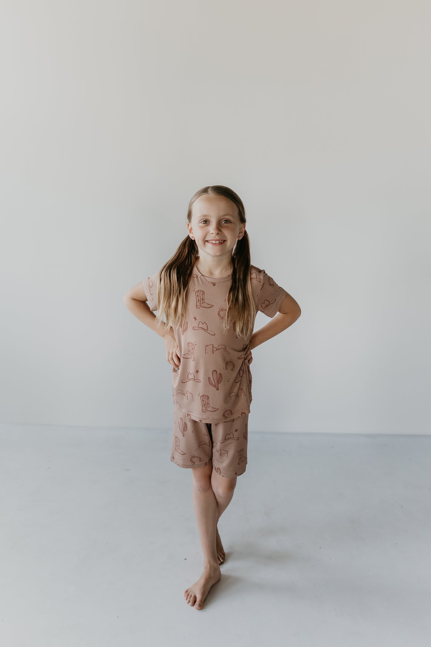 A young girl with long, light brown hair tied in pigtails stands barefoot against a plain white background. She is smiling and wearing the Bamboo Short Set from Minty x FF Wild West Desert Dreams, made from eco-friendly bamboo material featuring small, playful patterns. Her hands rest on her hips and one leg is slightly forward.