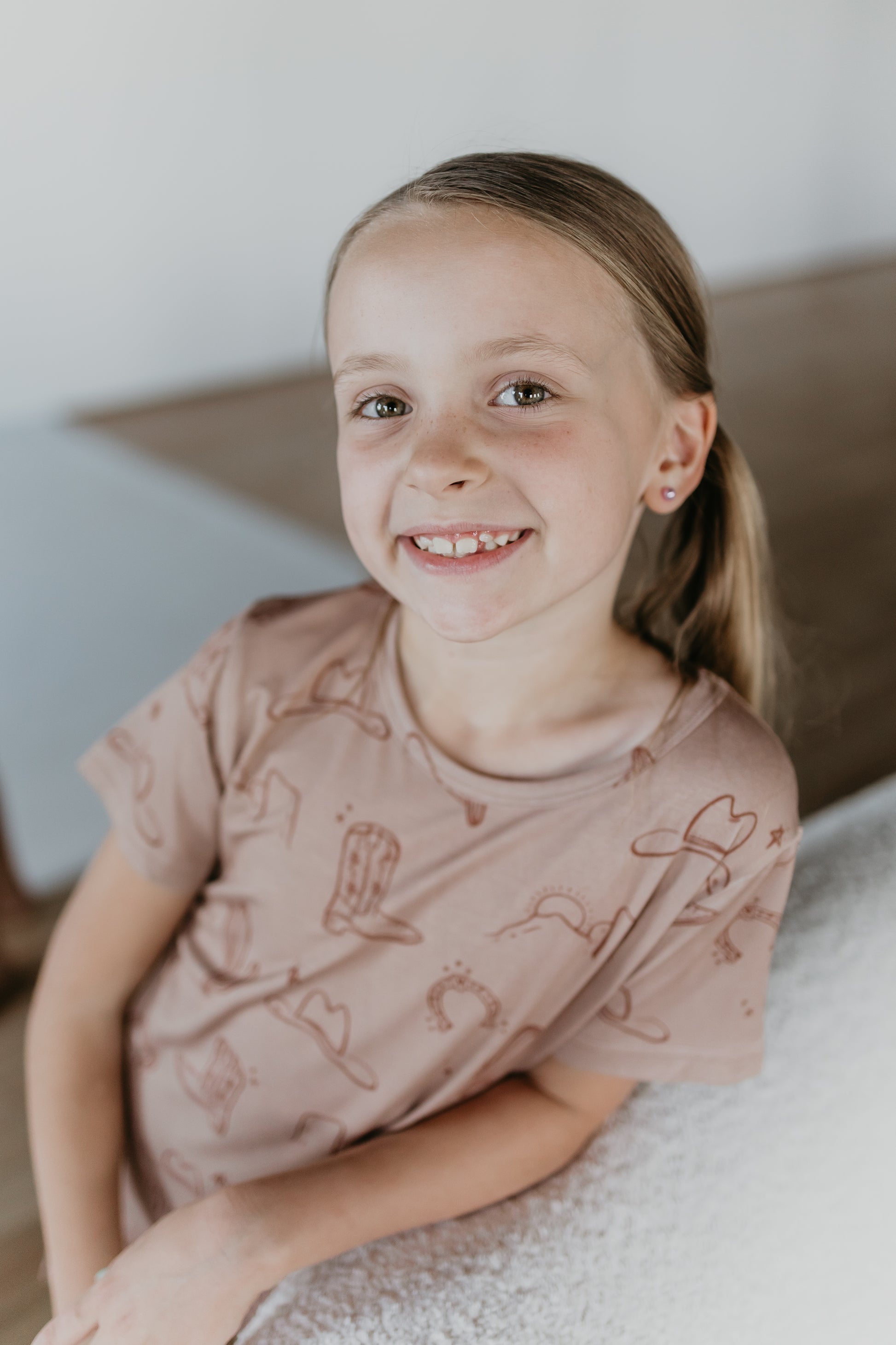 A young girl with blonde hair tied in a ponytail smiles at the camera. She is wearing the Bamboo Short Set from Minty x FF Wild West Desert Dreams, featuring a tan shirt with a pattern of cowboy hats, boots, and horseshoes made from eco-friendly bamboo material. She is leaning on a white surface with a light brown background.