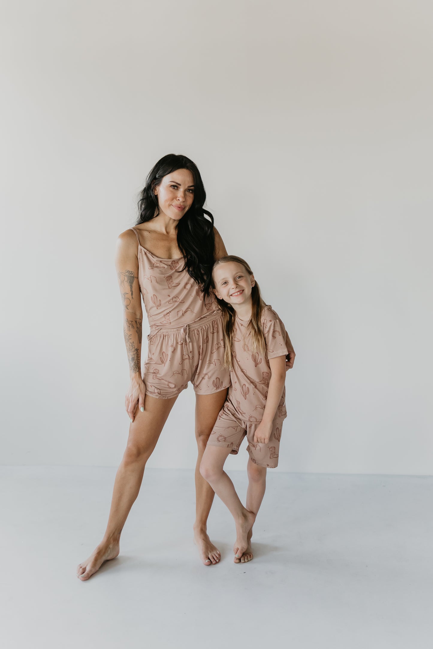 A woman with long dark hair and tattoos on her arms stands with a young girl with long blonde hair. They are both smiling, barefoot, and wearing matching light brown Bamboo Short Sets from the Minty x FF Wild West Desert Dreams collection, made from eco-friendly bamboo material featuring a minimalist pattern. The background is plain white.