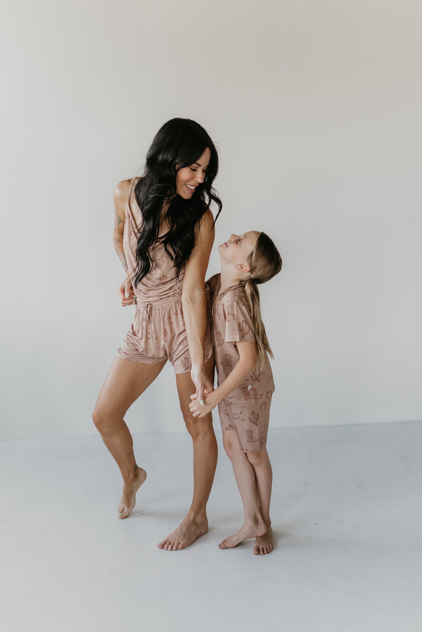 A woman and a young girl, both with long, dark hair, are standing in a minimalistic room. They are wearing matching beige Bamboo Short Sets from the Minty x FF Wild West Desert Dreams collection, made from eco-friendly bamboo material with playful designs. The hypo-allergenic outfits showcase their bond as the woman looks down at the smiling girl holding her hand.