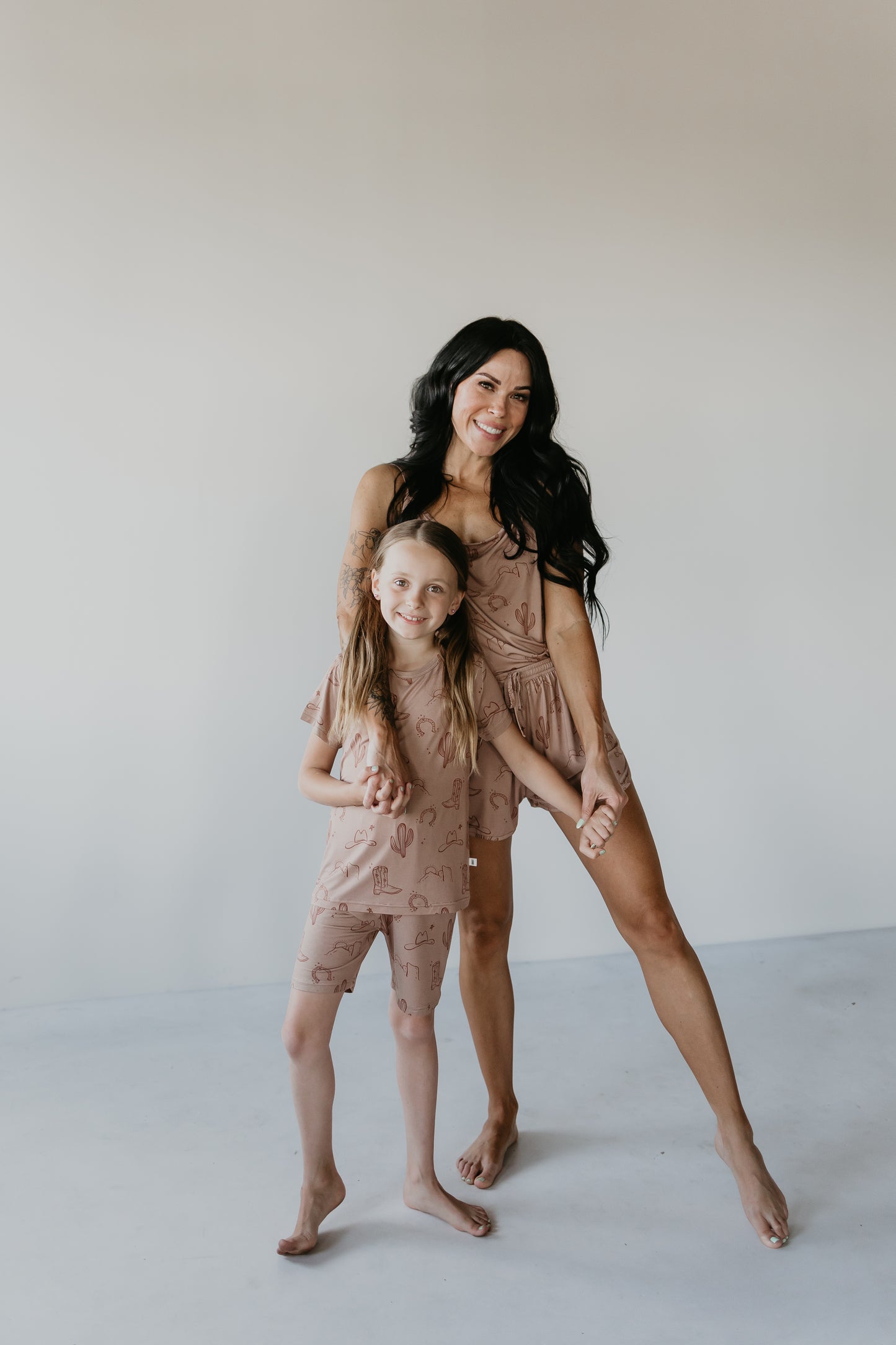 A smiling woman with long dark hair and tattoos on her arm holds hands with a young girl who has long blonde hair. They are both barefoot and wearing matching Bamboo Short Sets from the Minty x FF Wild West Desert Dreams collection, standing on a white floor against a plain light-colored wall.