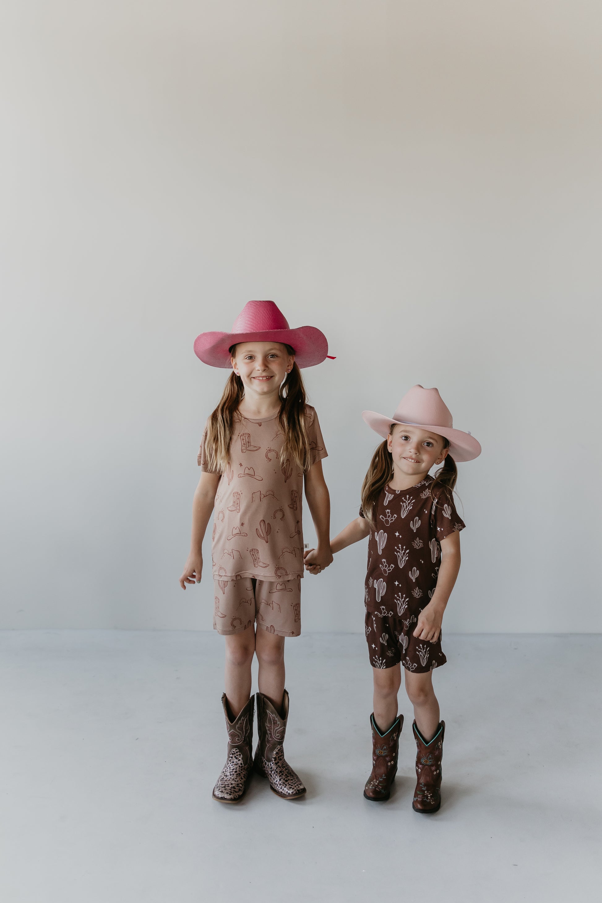 Two young girls are standing and holding hands against a plain background. They are both wearing cowboy boots and hats. The girl on the left has a pink hat and matching outfit, while the girl on the right sports eco-friendly Bamboo Short Set by Minty x FF Wild West Desert Dreams with a light pink hat and brown outfit featuring cactus prints.