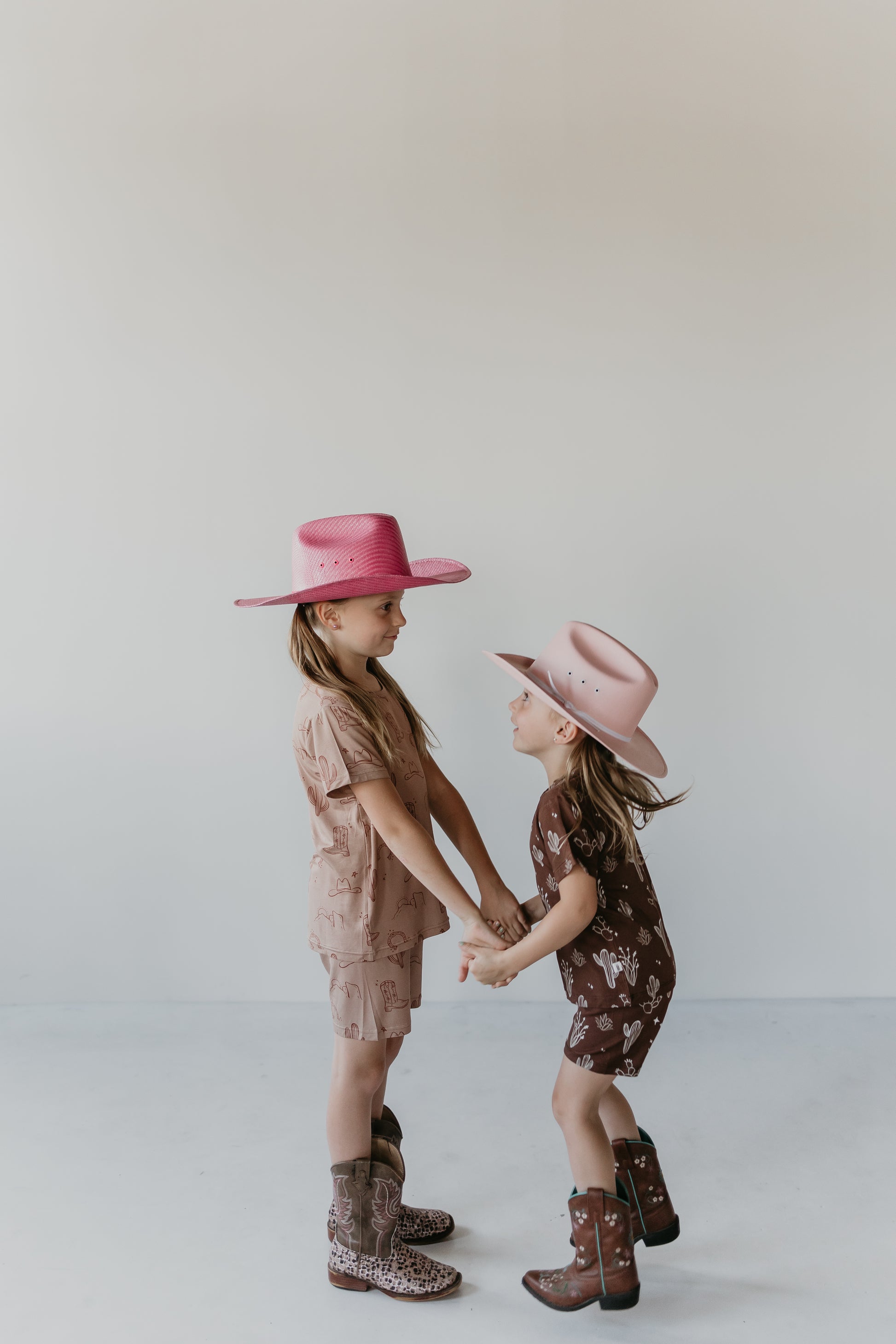 Two young girls wearing cowboy hats and boots stand facing each other, holding hands. Both are dressed in Minty x ff Wild West Desert Dreams Bamboo Short Sets—one in pink and the other in brown. Their light hair frames their happy, playful faces as they enjoy the comfort of eco-friendly bamboo material against a plain white wall.