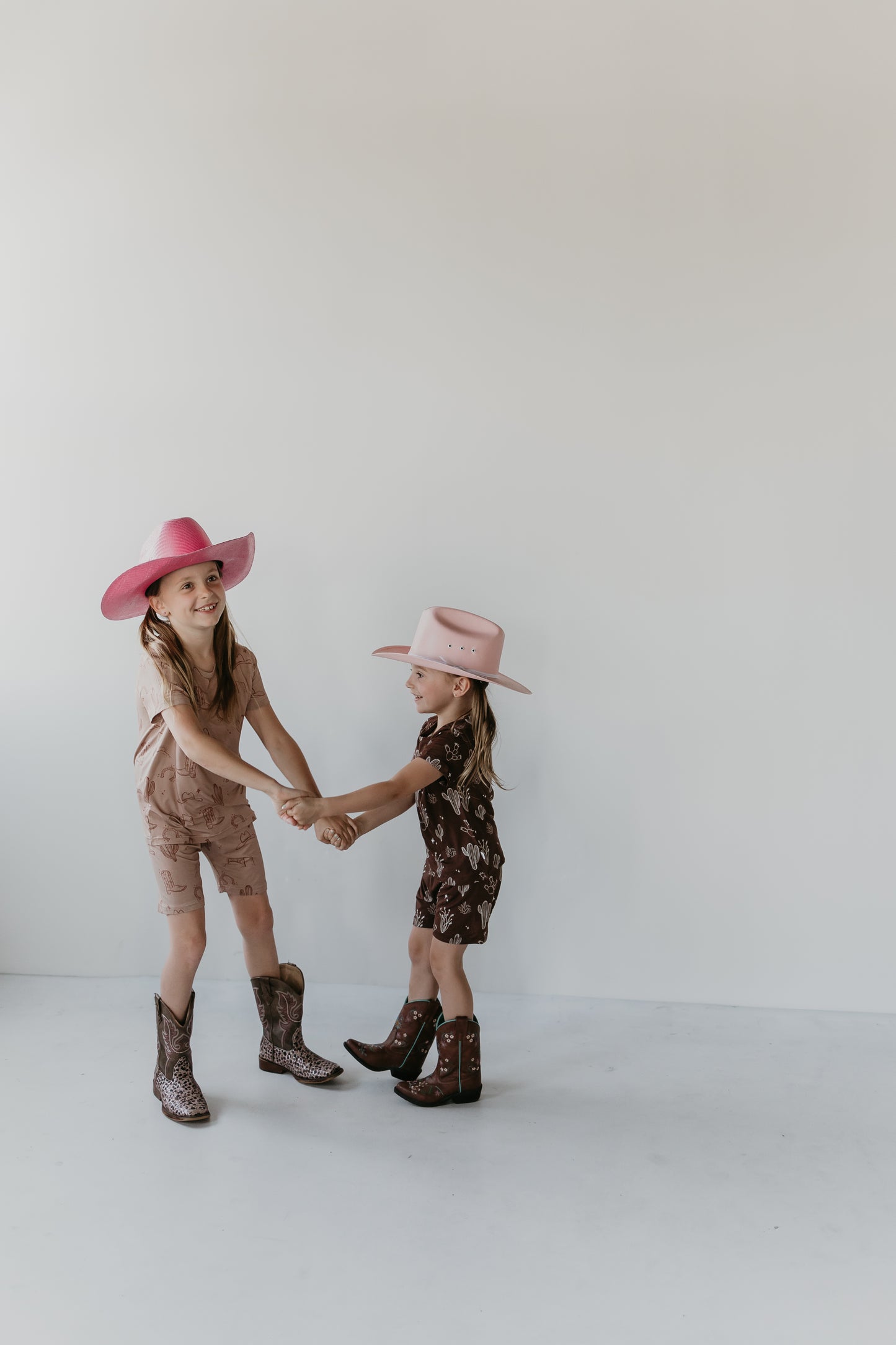 Two young girls in Minty x FF Wild West Desert Dreams' eco-friendly bamboo outfits and cowboy hats are holding hands and smiling in a minimalist, white, indoor setting. One girl wears a pink hat with a beige outfit, while the other sports a beige hat with a dark brown outfit. They seem to be dancing or playing together in their Bamboo Short Sets from the Minty x ff Wild West collection.