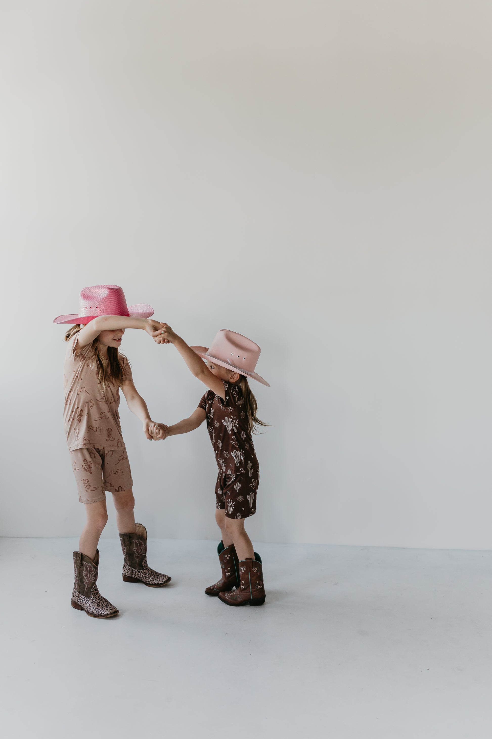 Two young children wearing cowboy hats and boots are holding hands and smiling. One child wears a pink hat and a light brown Bamboo Short Set from the Minty x FF Wild West Desert Dreams collection, while the other wears a white hat and dark brown outfit. They dance playfully against a simple white background.