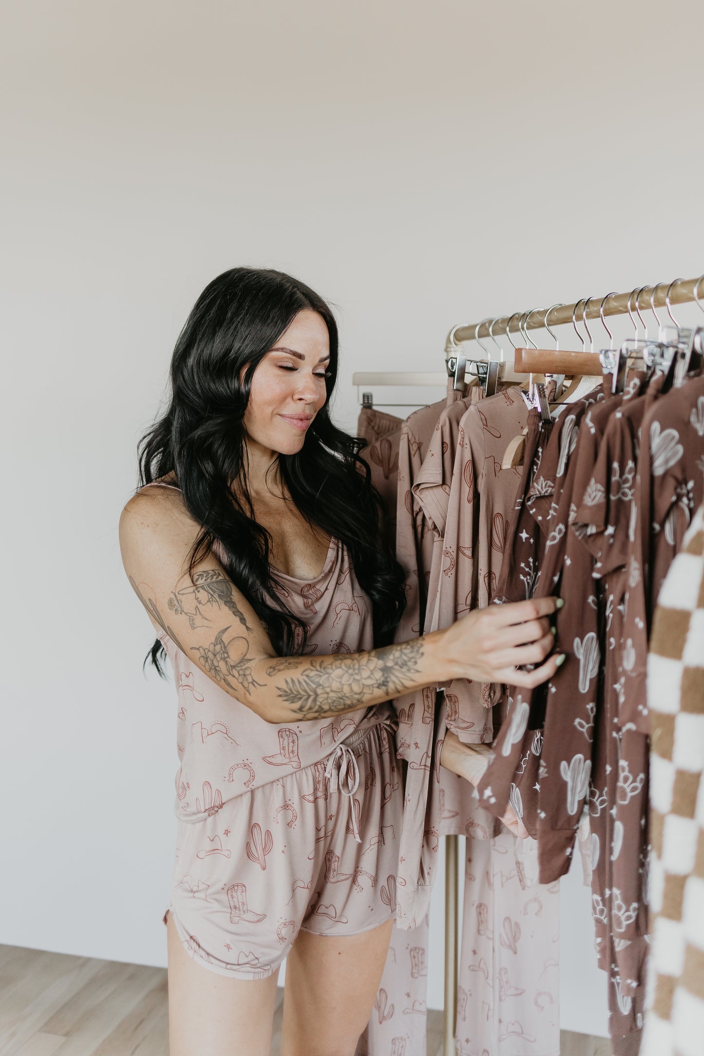 A woman with long dark hair and tattoos on her arms is wearing the Bamboo Short Set from Minty x FF Wild West's Desert Dreams collection. She is smiling while looking at and touching clothes hanging on a rack, which include various patterned garments made of eco-friendly bamboo material.