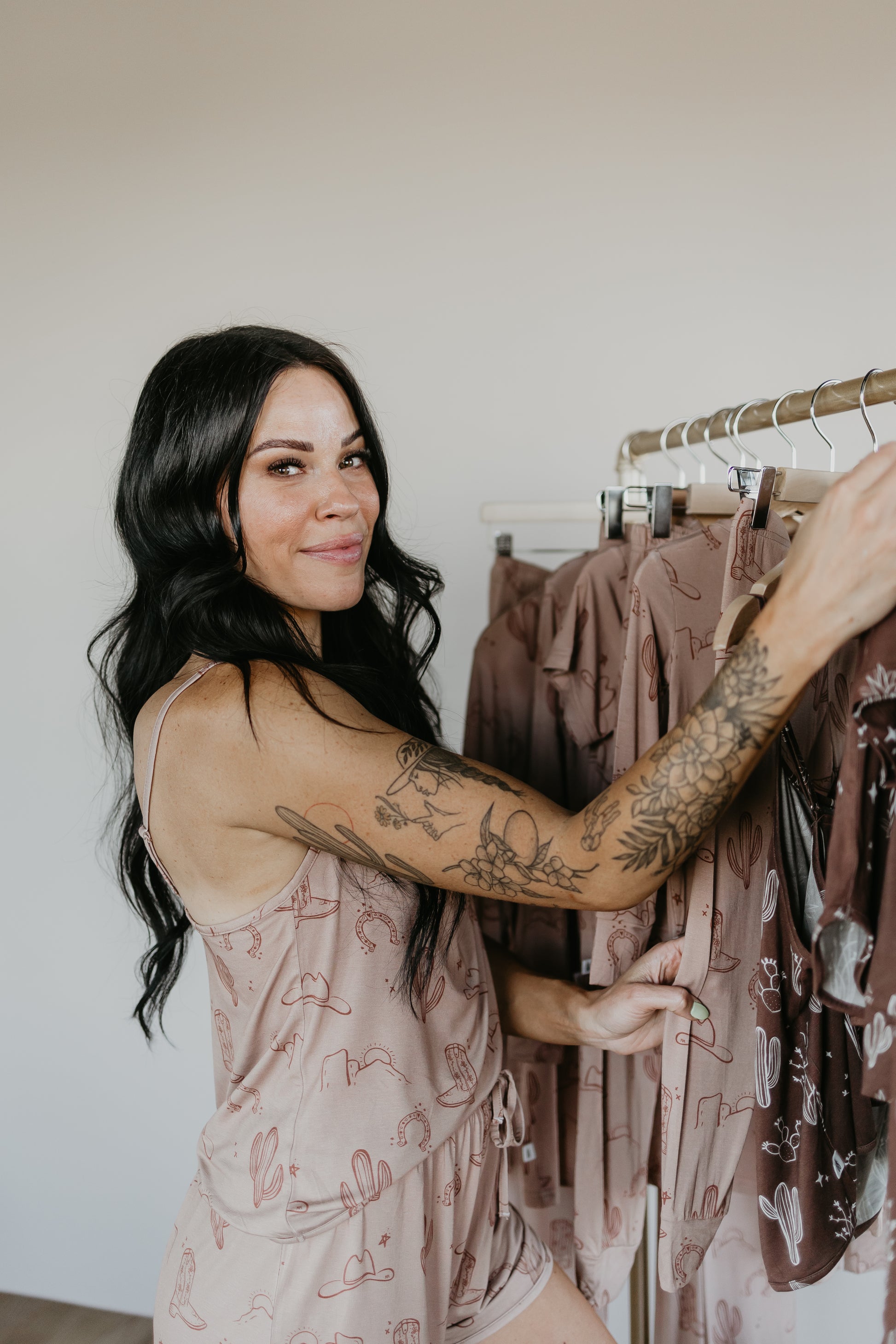 A person with long dark hair and tattoos stands beside a clothing rack, wearing the Minty x ff Desert Dreams Bamboo Two Piece Pajamas decorated with a cactus pattern. They're smiling warmly at the camera against a neutral background, perfectly embodying the Minty x FF Wild West Desert Dreams vibe.