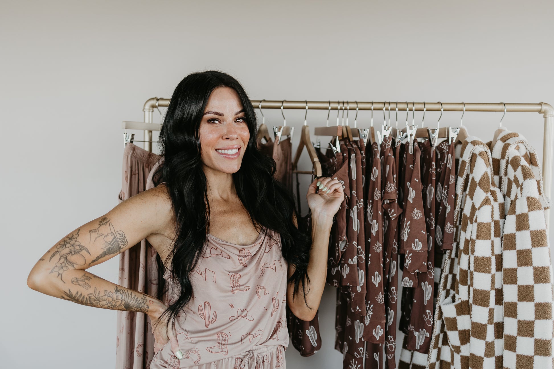 A smiling person with long dark hair stands in front of a clothing rack, wearing an Adult Hooded Robe from the Minty x FF Wild West Desert Dreams collection. The rack features items from Forever French Baby, showcasing earthy tones and patterns made from luxurious microfiber feather yarn.