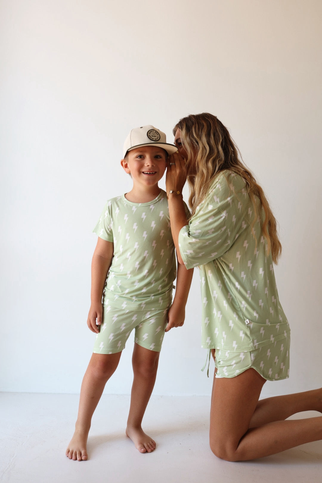 A woman kneels beside a smiling boy, whispering in his ear. Both wear the forever french baby Bamboo Short Set | Flash, featuring light green bamboo fabric with white lightning bolts. The boy also wears a white cap in a bright, minimal room.