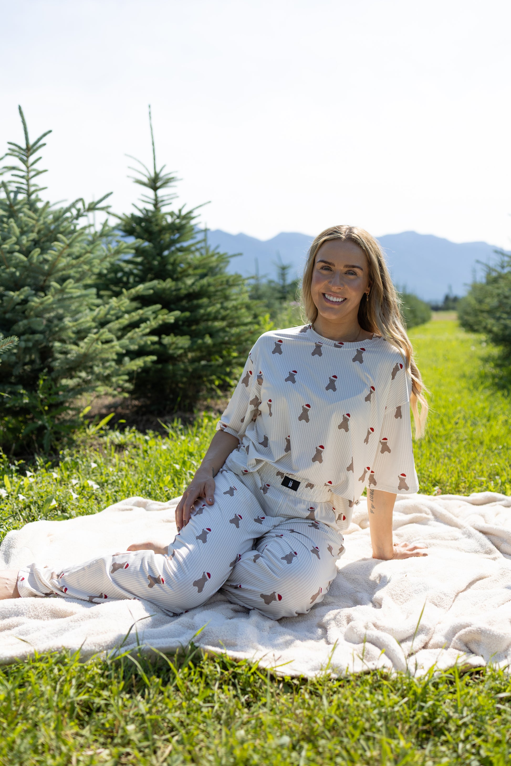 A woman sits on a blanket in a grassy field near pine trees and mountains, smiling at the camera. She wears the Women's Boxy Tee Lounge Set | Santa Teddy by forever french baby—a breathable loungewear set featuring a playful bear print. The scene is bright and sunny.