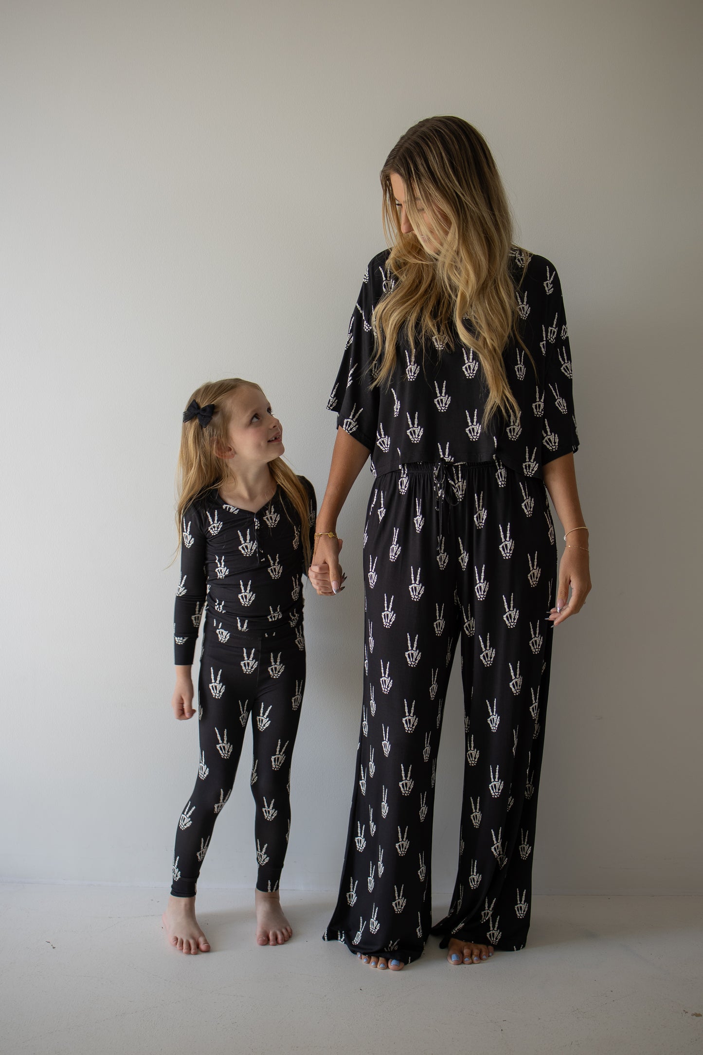 A woman and young girl stand barefoot against a plain wall, holding hands and smiling. They wear matching forever french baby Bamboo Two Piece Pajamas in the Midnight Skelly print—ultra-soft, breathable bamboo clothing that’s perfect for sensitive skin.