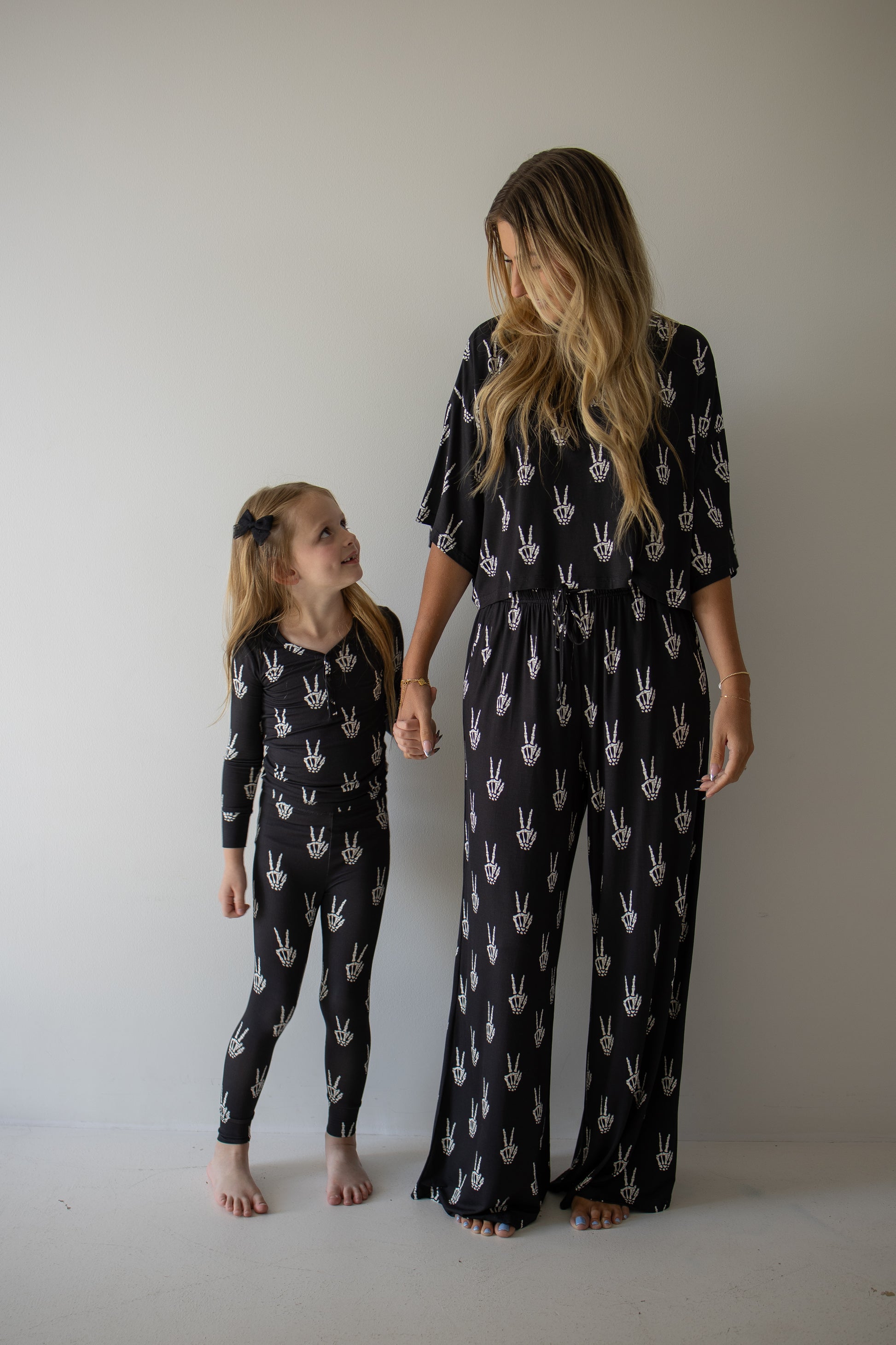 A woman and young girl stand barefoot against a plain wall, holding hands and smiling. They wear matching forever french baby Bamboo Two Piece Pajamas in the Midnight Skelly print—ultra-soft, breathable bamboo clothing that’s perfect for sensitive skin.