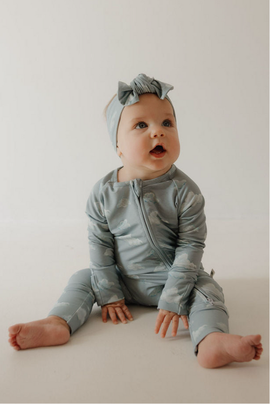 A baby in Kendy 3’s Bamboo Zip Pajamas | Duke, paired with a matching bow headband, sits on the floor and looks up cheerfully. The hypo-allergenic pajamas provide year-round comfort against a plain, light backdrop.