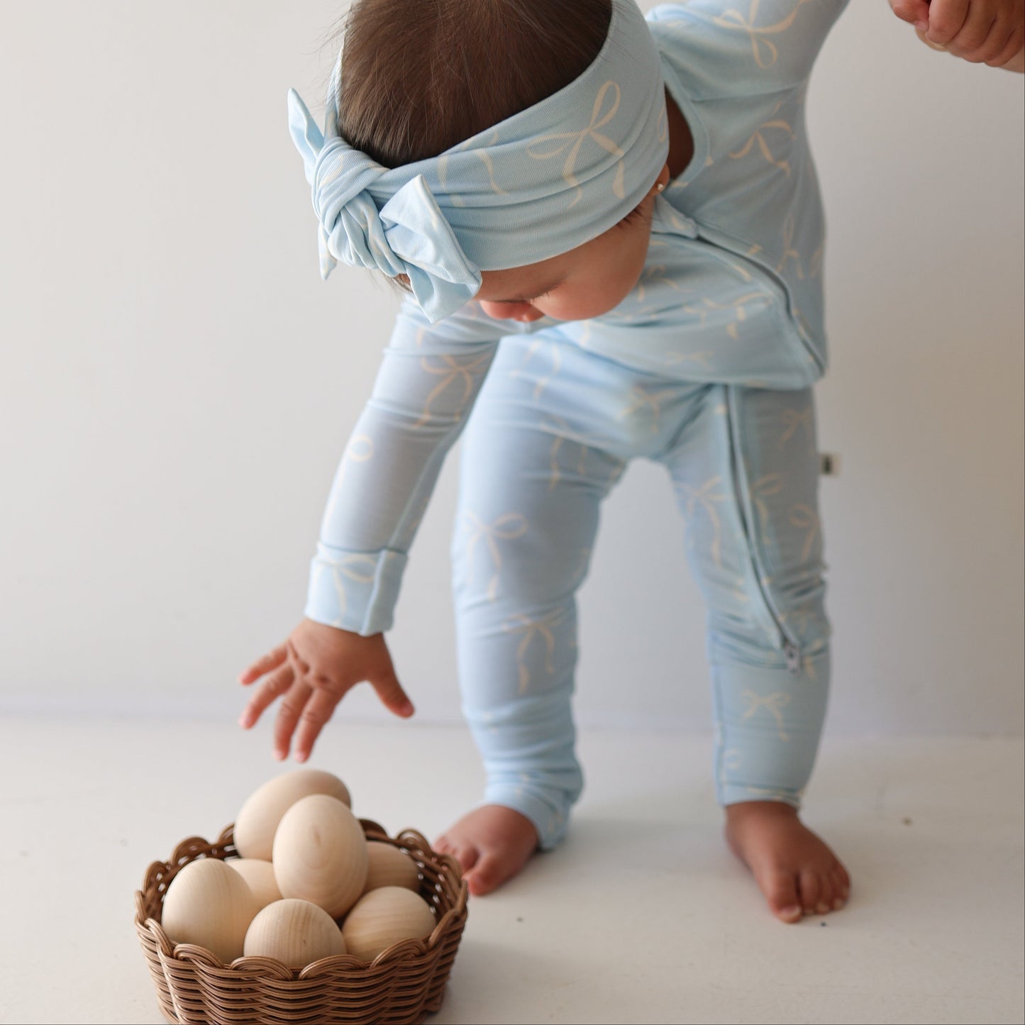 A young child wearing forever french baby Bamboo Zip Pajamas in Blue Bows with a matching headband reaches for a basket of wooden eggs on a white floor.