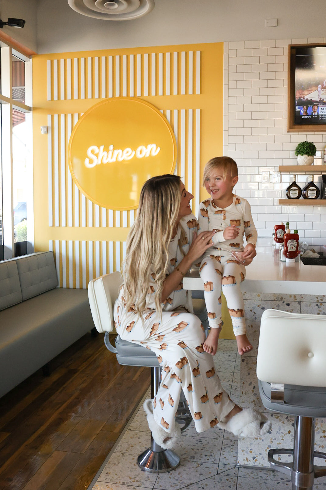 A woman and young child wear matching "Sunday Stack" Bamboo Two Piece Pajamas by forever french baby, featuring playful dog prints as they smile at a diner counter. A bright yellow "Shine on" sign adds more warmth to their cozy moment together.