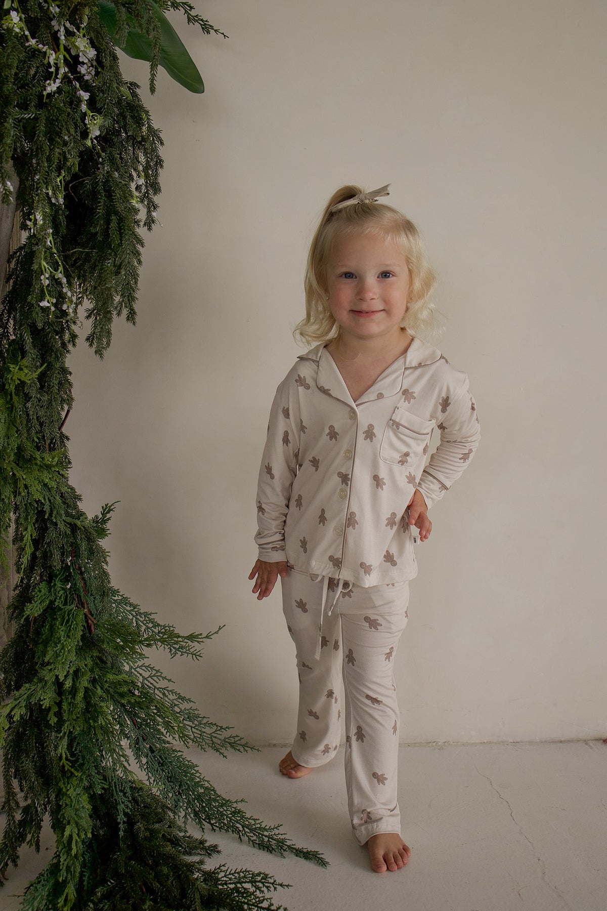 A toddler wearing a Girl’s Bamboo Button Up Lounge Set | Gingersnap featuring a long-sleeved button-up shirt and matching pants decorated with a repeating gingerbread man pattern.