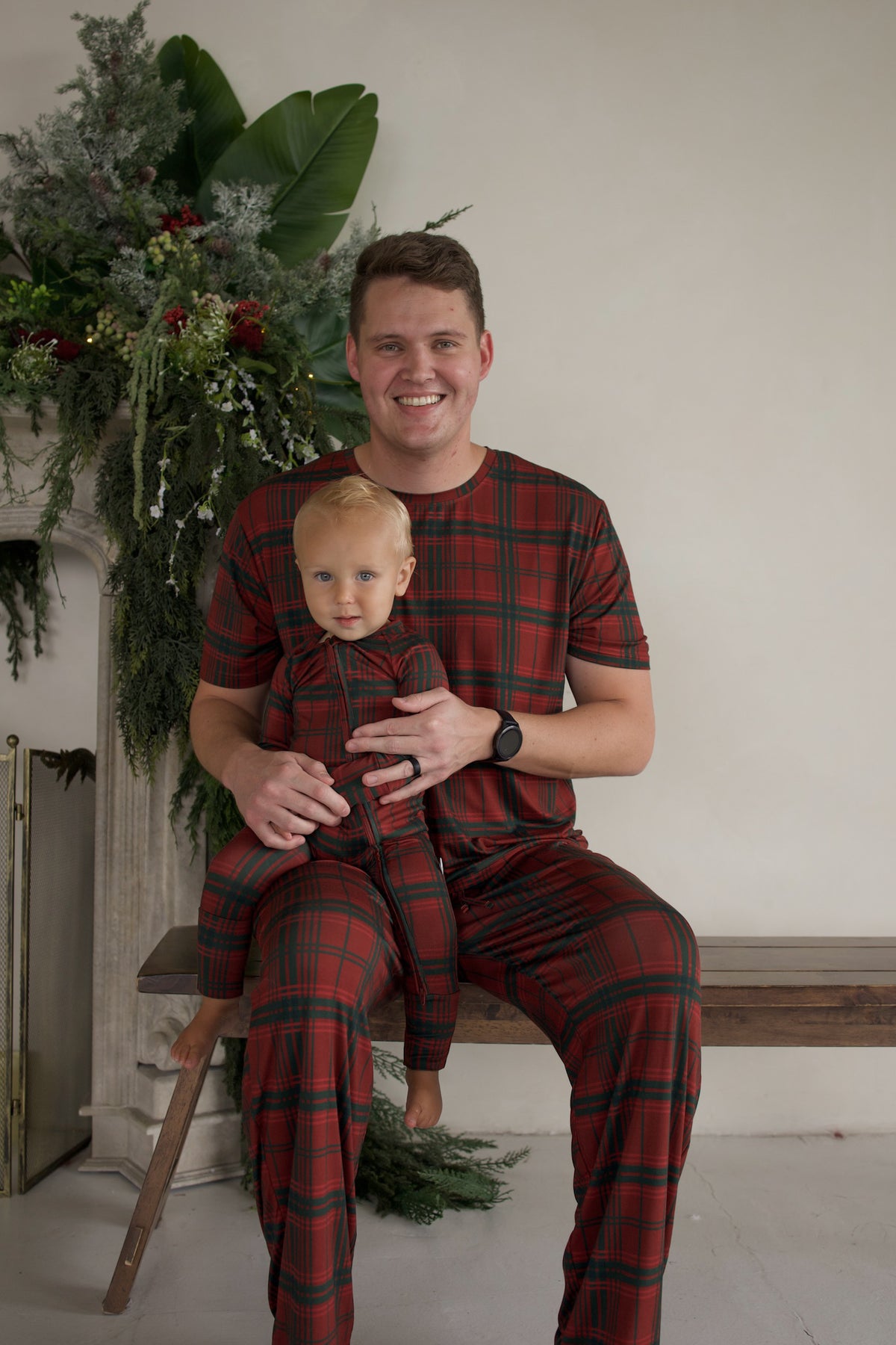 A man sitting on a wooden bench holds a toddler on his lap, both wearing matching Men’s Short Sleeve Pajamas | Christmas Plaid by forever french baby.
