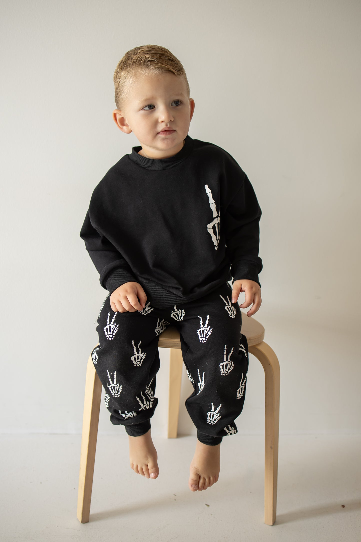 A young child sits barefoot on a wooden stool, dressed in the forever french baby Child Sweat Set | Midnight Skelly—featuring striking white skeleton hand designs on both the sweatshirt and matching pants—set against a plain light background, making it perfect for everyday adventures.