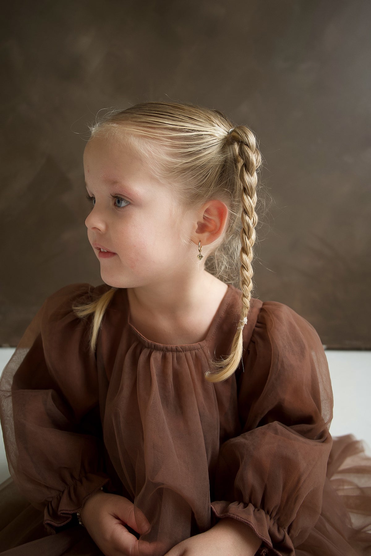 A young girl with blonde hair in a braid, wearing the Long Sleeve Tutu Dress | Cocoa by forever french baby, sits and gazes to the side against a brown backdrop.