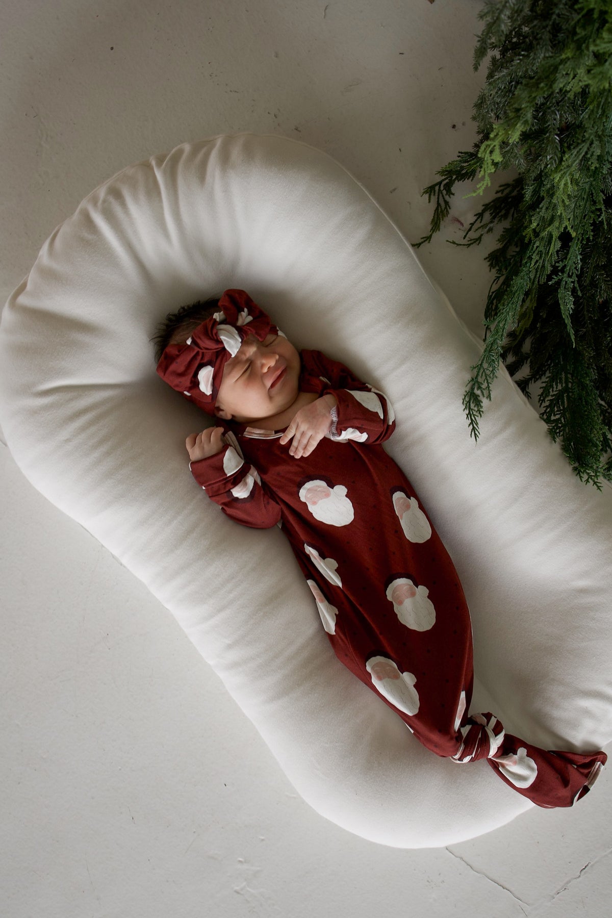 A newborn baby wearing a Bamboo Knotted Gown | Vintage Santa and matching headwrap, lying in a white baby lounger.