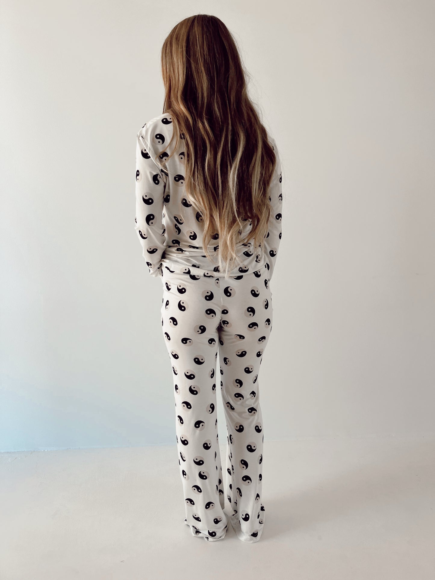 A person with long, wavy hair stands facing away from the camera, wearing a matching set of Women's Bamboo Pajamas in black and white adorned with yin-yang symbols from the brand forever French baby. The background is a plain, light-colored wall.