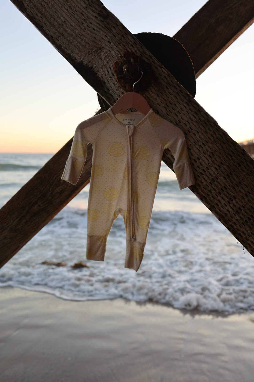 The forever french baby Bamboo Zip Pajamas | Lemon Drop, a small yellow-patterned hypo-allergenic onesie, hangs on a wooden beam by the beach with waves and a sunset in the background.
